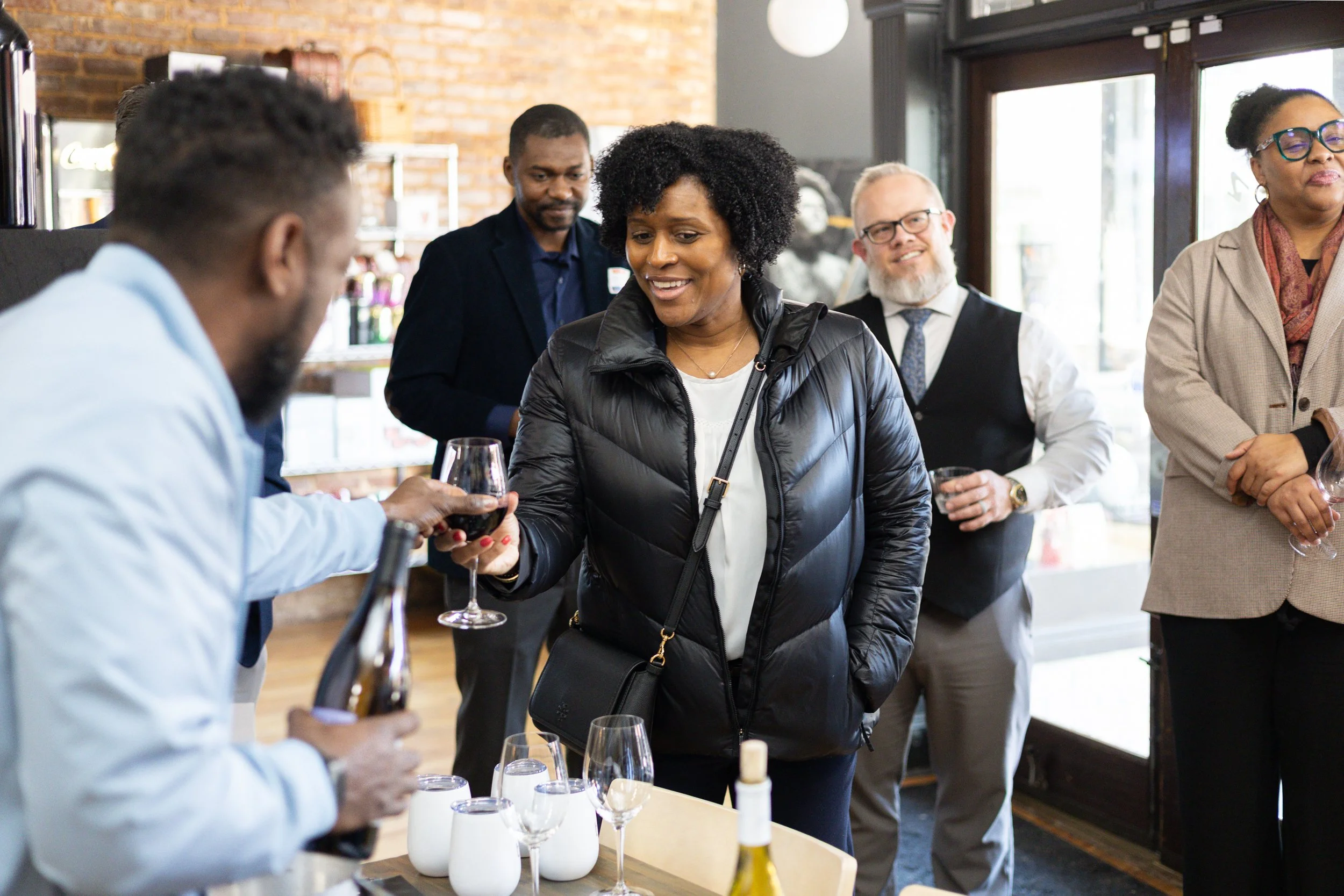 A woman in a black jacket receives a glass of red wine from a man in a white shirt at a social gathering in a restaurant or cafe.