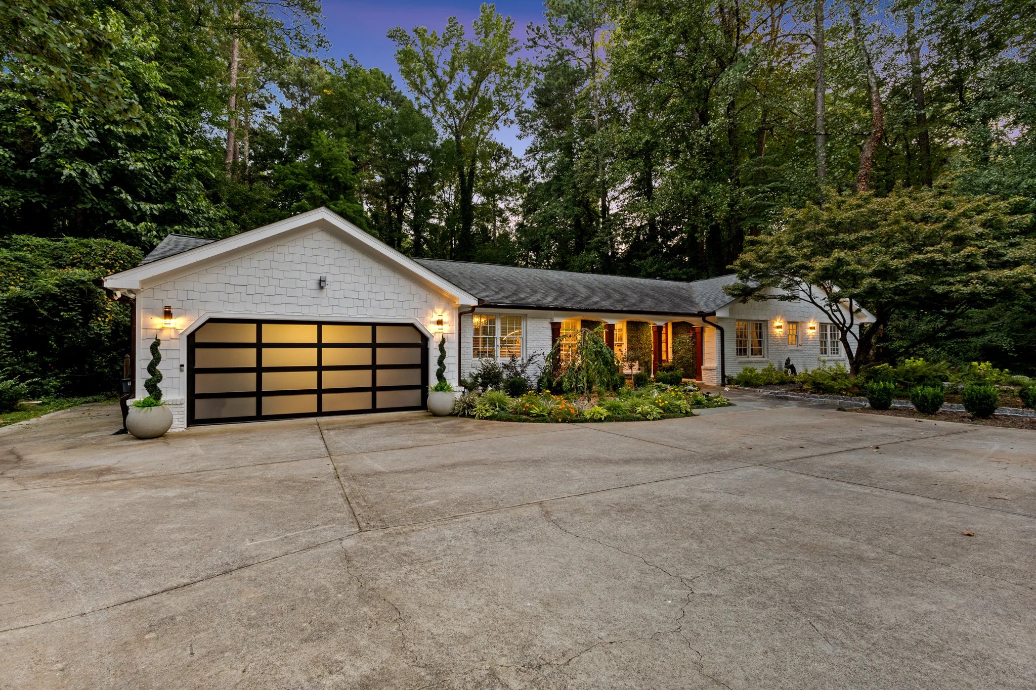 A white brick house with a black and frosted glass garage door, surrounded by trees and landscaped garden, illuminated by exterior wall lights at dusk.