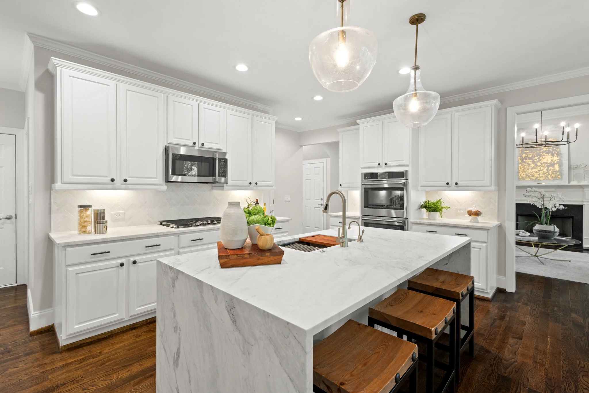 Modern white kitchen with marble island, wooden stools, stainless steel appliances, and pendant lights.