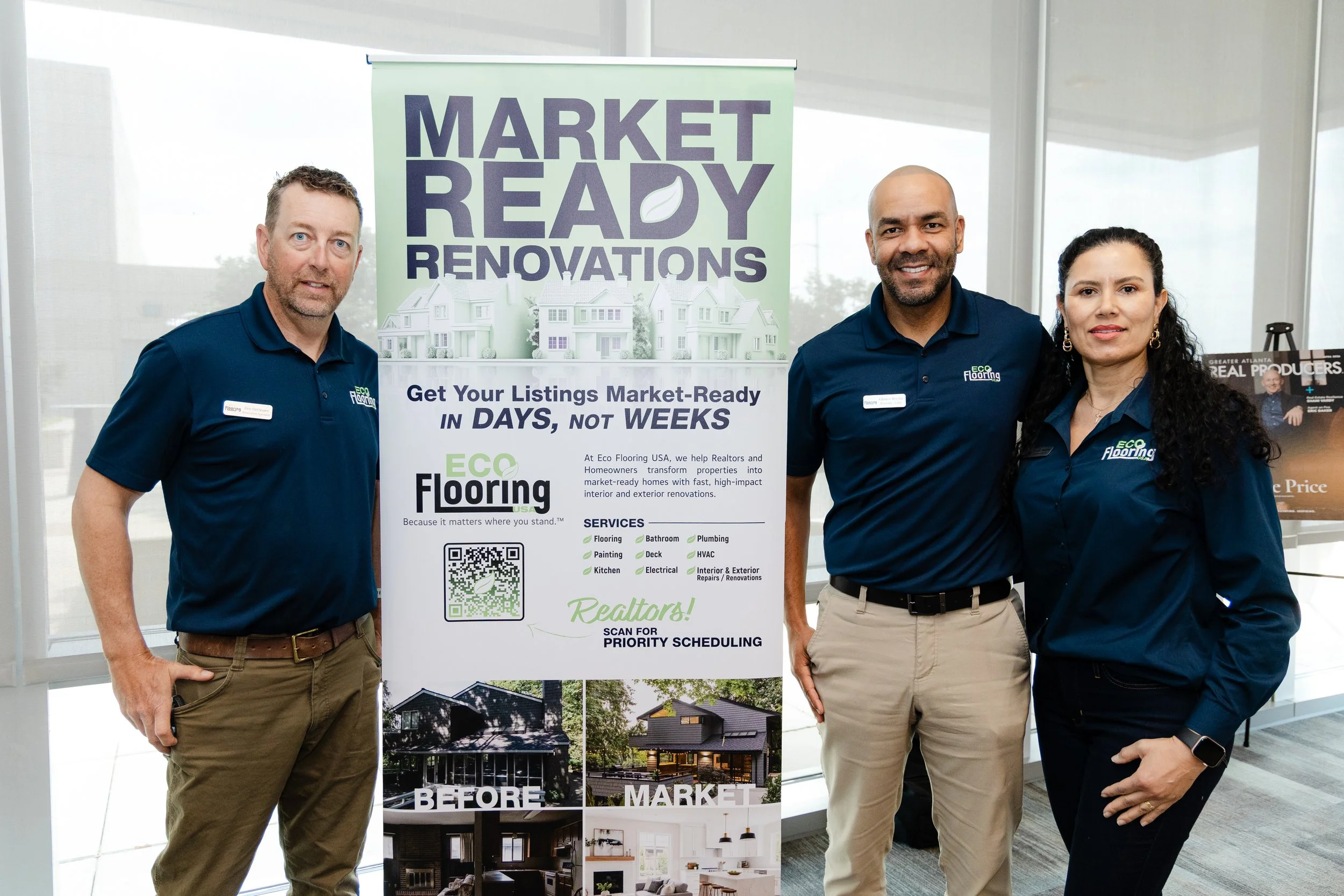Three people standing in front of a green and white banner for Eco Flooring USA, with images of house renovations below them. They are indoors, wearing matching blue shirts with the company's logo, and are smiling. The man on the left has short hair and a beard, the man in the middle is bald with a beard, and the woman on the right has long dark curly hair.