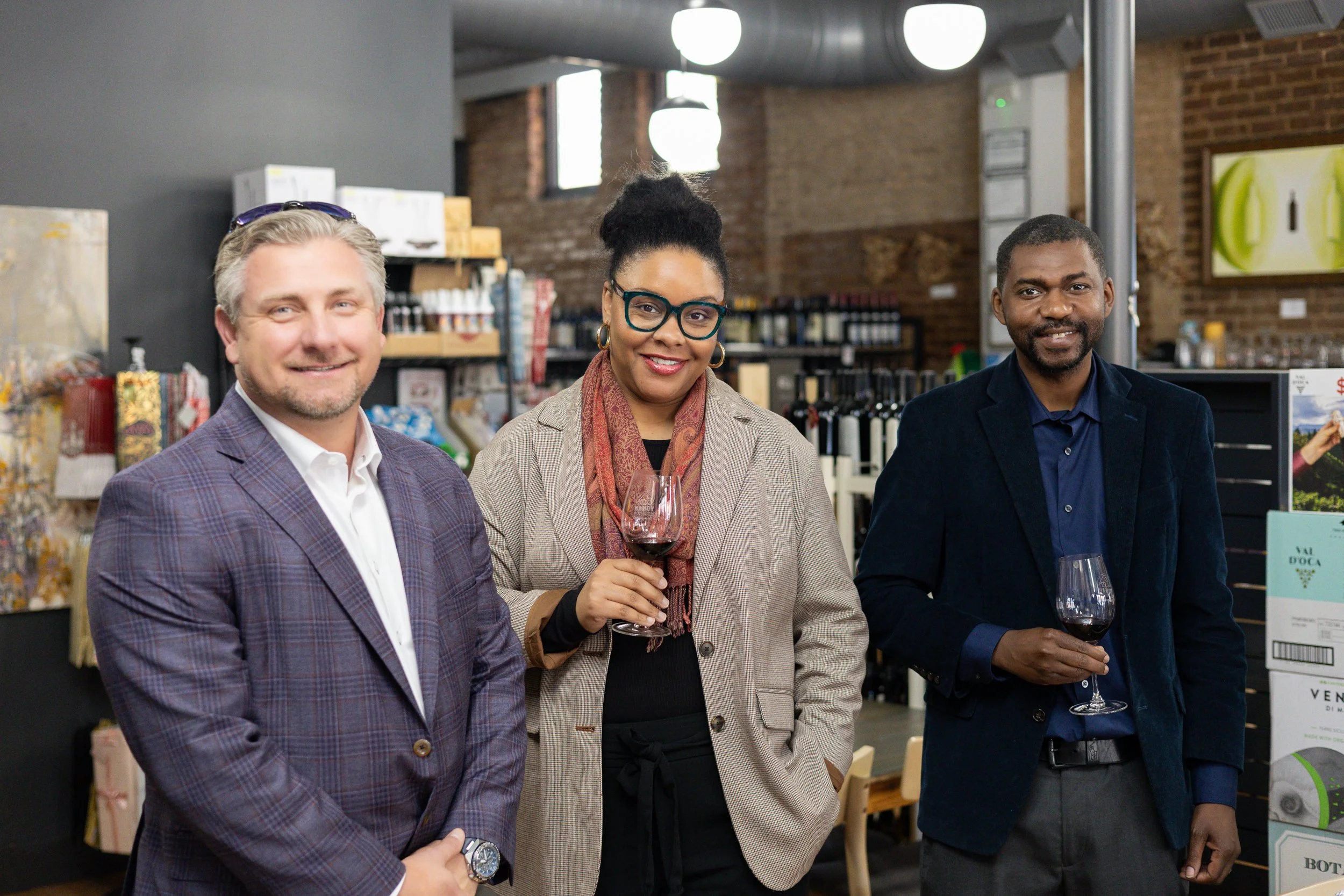 Three diverse people with glasses of red wine in a store or wine shop, standing in line and smiling at the camera.