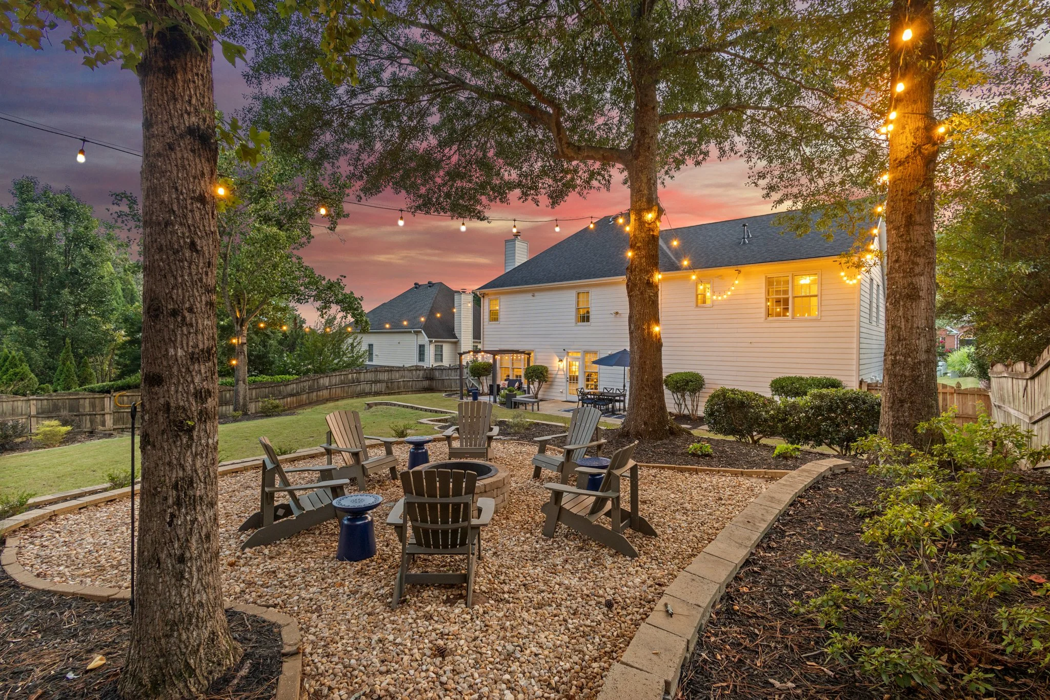 Backyard patio with Adirondack chairs arranged in a circle around a fire pit, string lights hanging from trees, and a white house in the background during sunset.