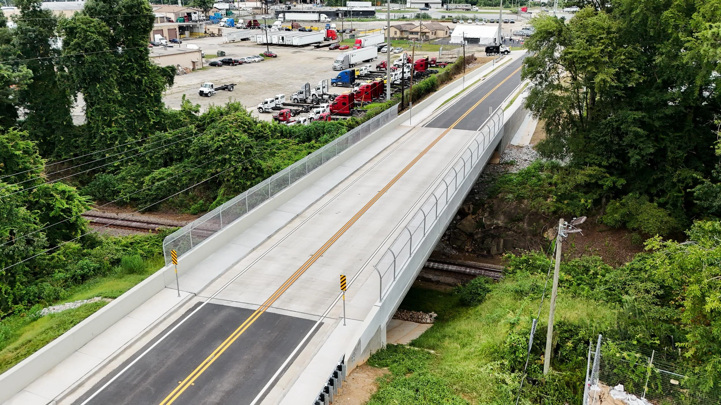 A newly constructed concrete bridge for vehicles spans over a small creek, with trees and green foliage on both sides. Below the bridge are train tracks, and in the background, there is a parking lot filled with trucks and cars, and some buildings.