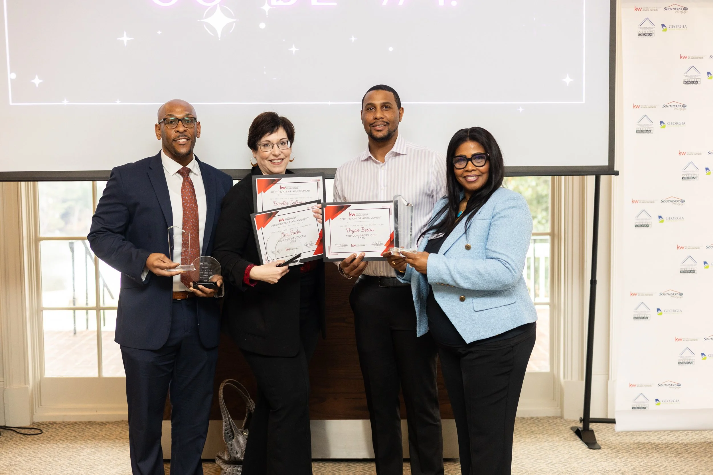 Four people holding awards and certificates, smiling at an award ceremony in a bright room with large windows and a projection screen behind them.