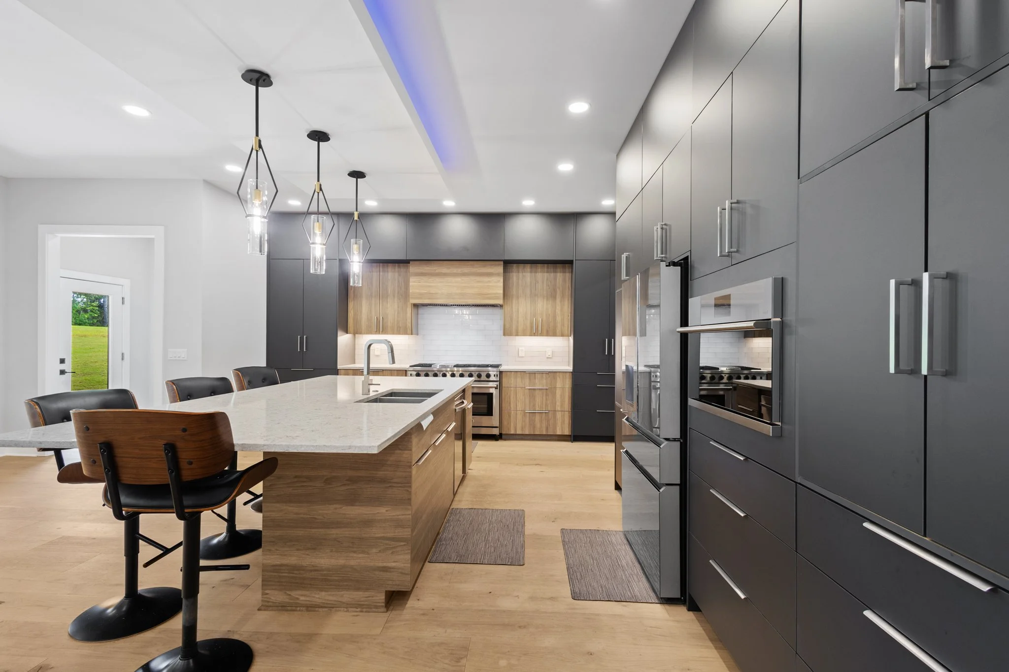 Modern kitchen with gray cabinets, a large island with a light-colored granite countertop, black barstools, and pendant lights, hardwood floors, and a door leading outside.
