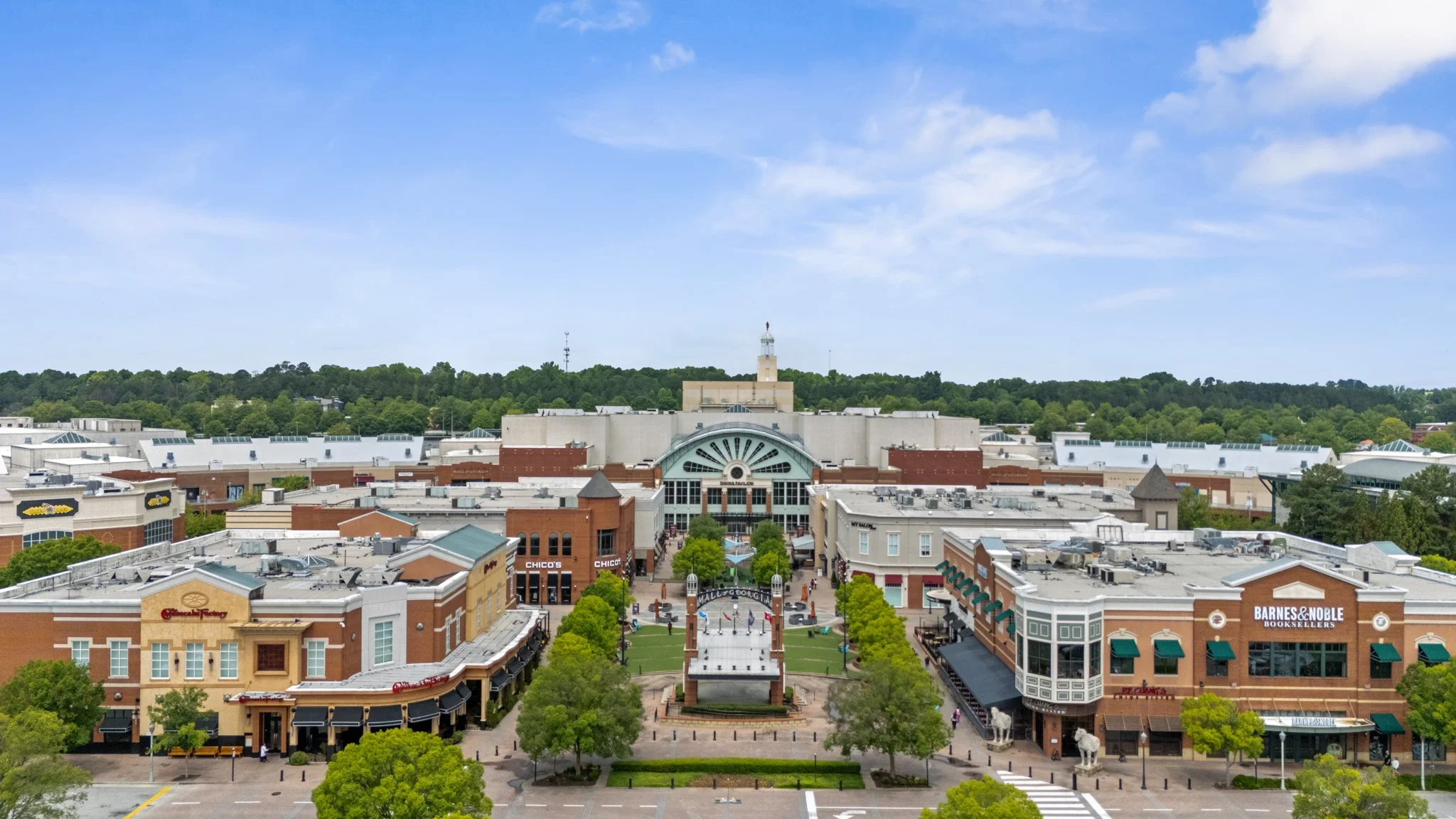 Aerial view of the Mall of Georgia in Buford, Georgia with a clock tower, surrounded by buildings and green trees under a partly cloudy sky.