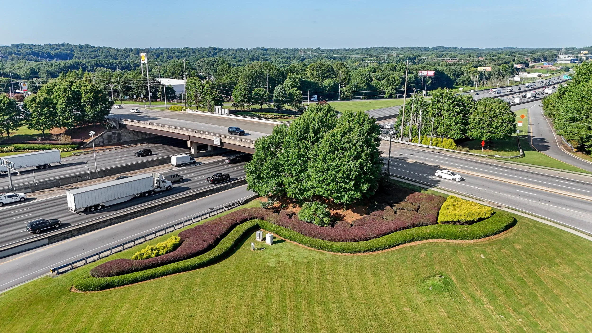 Aerial view of a multi-level highway interchange with cars and trucks, surrounded by green trees, grass, and landscaped areas. In the background, there are commercial buildings and a distant hilly landscape under a blue sky.