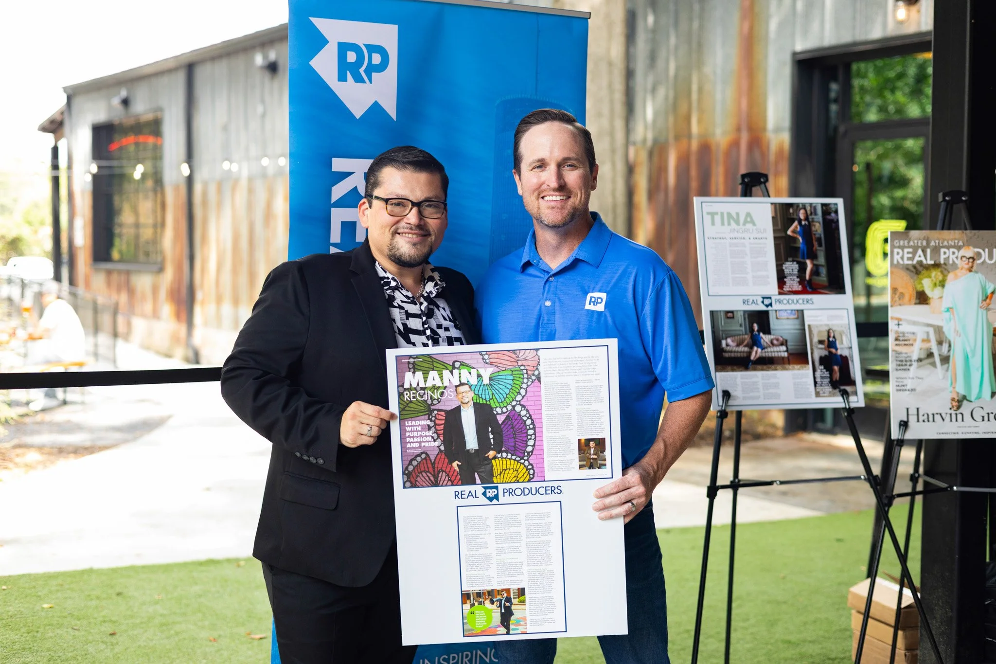 Two men standing side by side, holding a large poster with the title "Manny Reginos" and the logo "REAL RP PRODUCERS." One man is dressed in a black suit with a patterned shirt, and the other is in a blue polo shirt with the same logo. Behind them is a blue banner with the same logo, and display boards with articles and images are visible. The setting appears to be an outdoor or semi-outdoor event space with rustic metal siding and greenery.