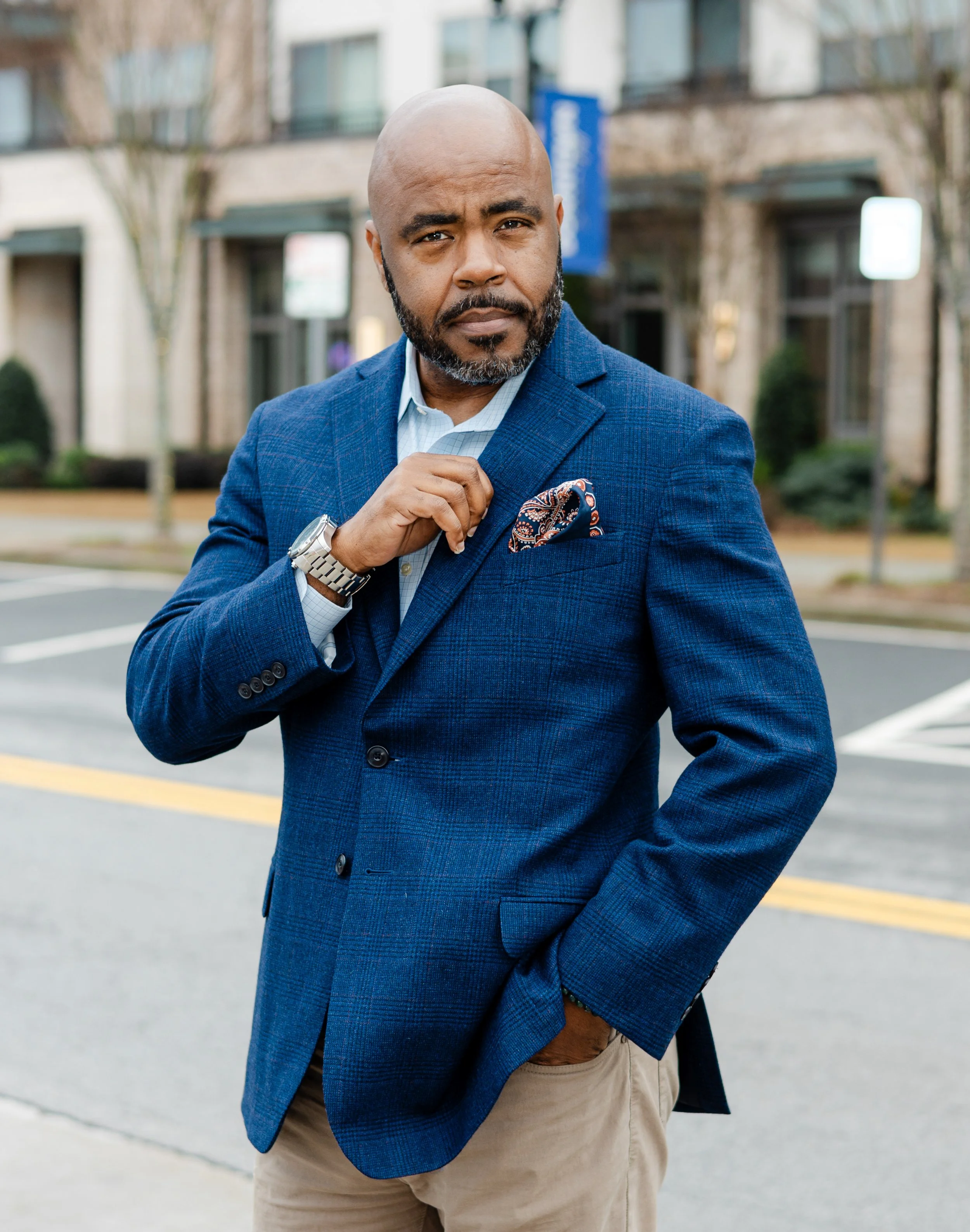 A confident man in a blue blazer and beige pants standing outdoors on a city street, adjusting his pocket square with a serious expression.