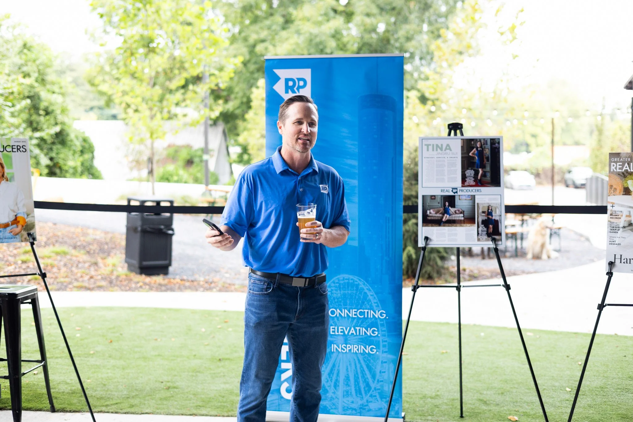 Man in blue shirt holding a drink and a phone, speaking at an outdoor event with display boards and trees in the background.
