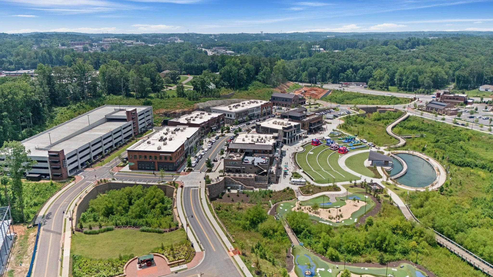 Aerial view of a modern shopping and entertainment development with multi-story parking garages, retail storefronts, landscaped walkways, green spaces, water features, and a surrounding wooded area and residential neighborhood.