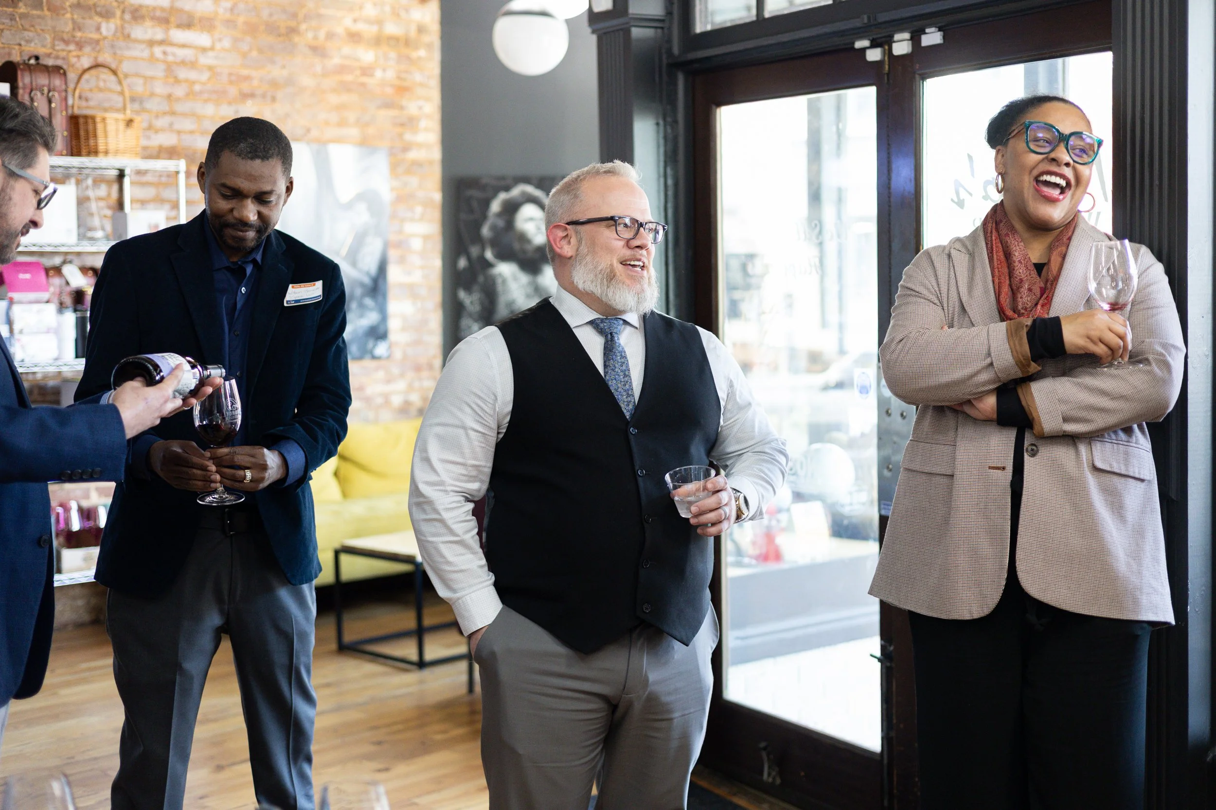 Group of four diverse adults socializing at a cafe, with one person pouring wine, two holding glasses, and one smiling and laughing.