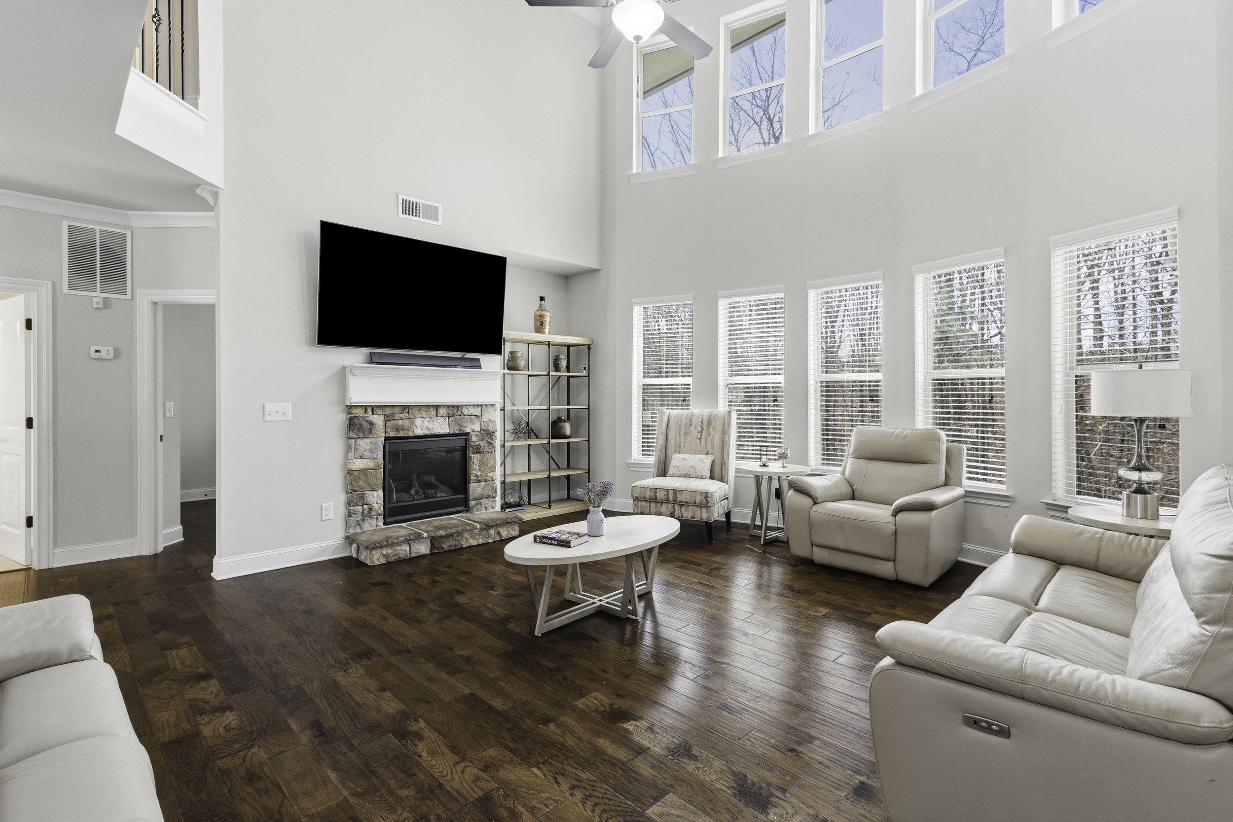 Bright living room with high ceiling, large windows, and a ceiling fan. White walls, hardwood floor, a stone fireplace, a wall-mounted TV above the fireplace, beige leather armchairs and sofas, a small round table between the chairs, a white coffee t