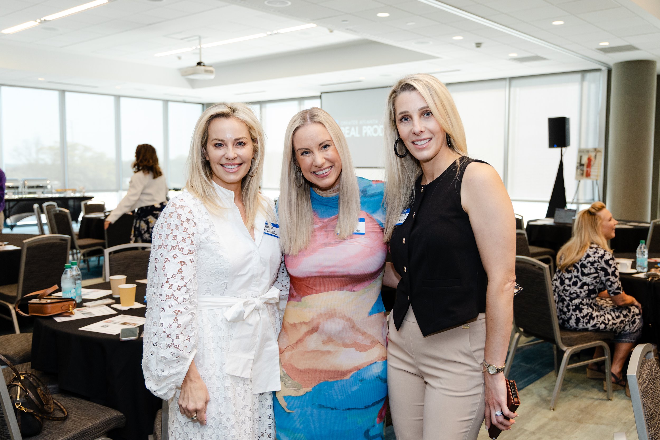 Three women smiling and standing together at a conference or event in a well-lit room with large windows and tables, with other attendees in the background.