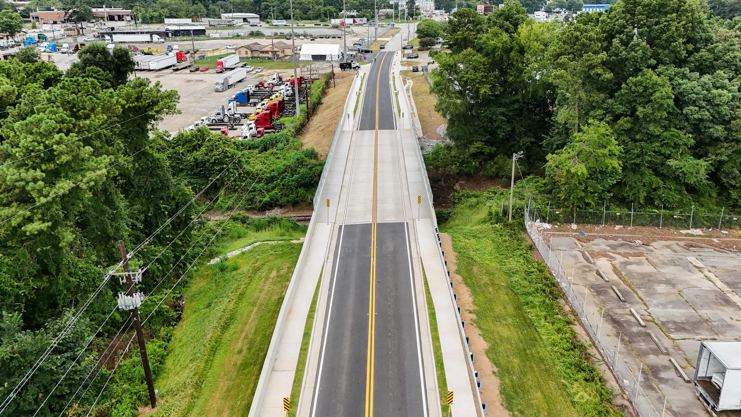 An aerial view of a small bridge crossing over a railway track, surrounded by dense trees on both sides. There are commercial and industrial areas visible beyond the bridge, with parked trucks, trailers, and industrial buildings.