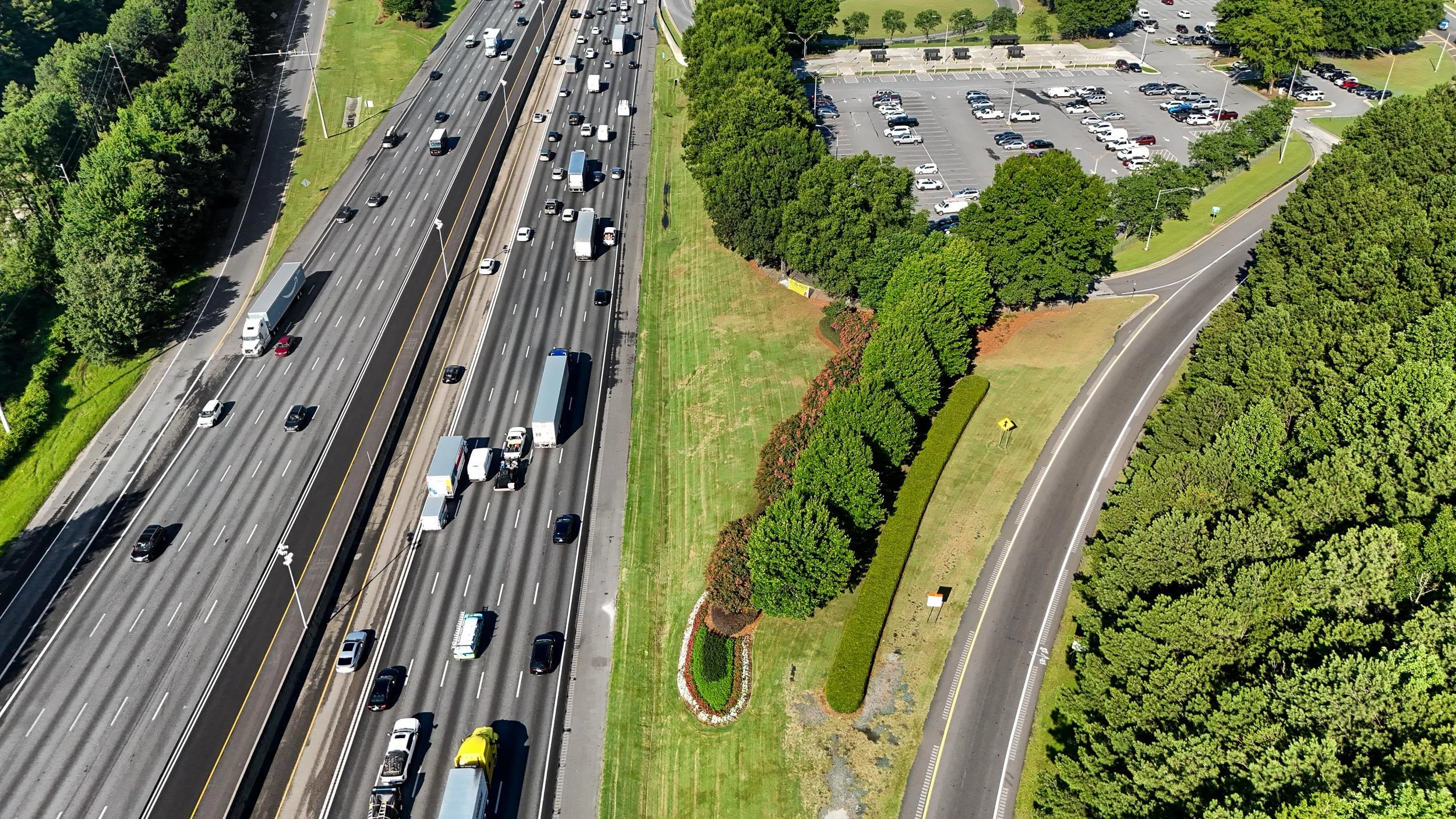 An aerial view of a busy Highway 85 South in Gwinnett County, Georgia with multiple lanes of cars and trucks, adjacent to a landscaped area with trees, grass, and a parking lot.