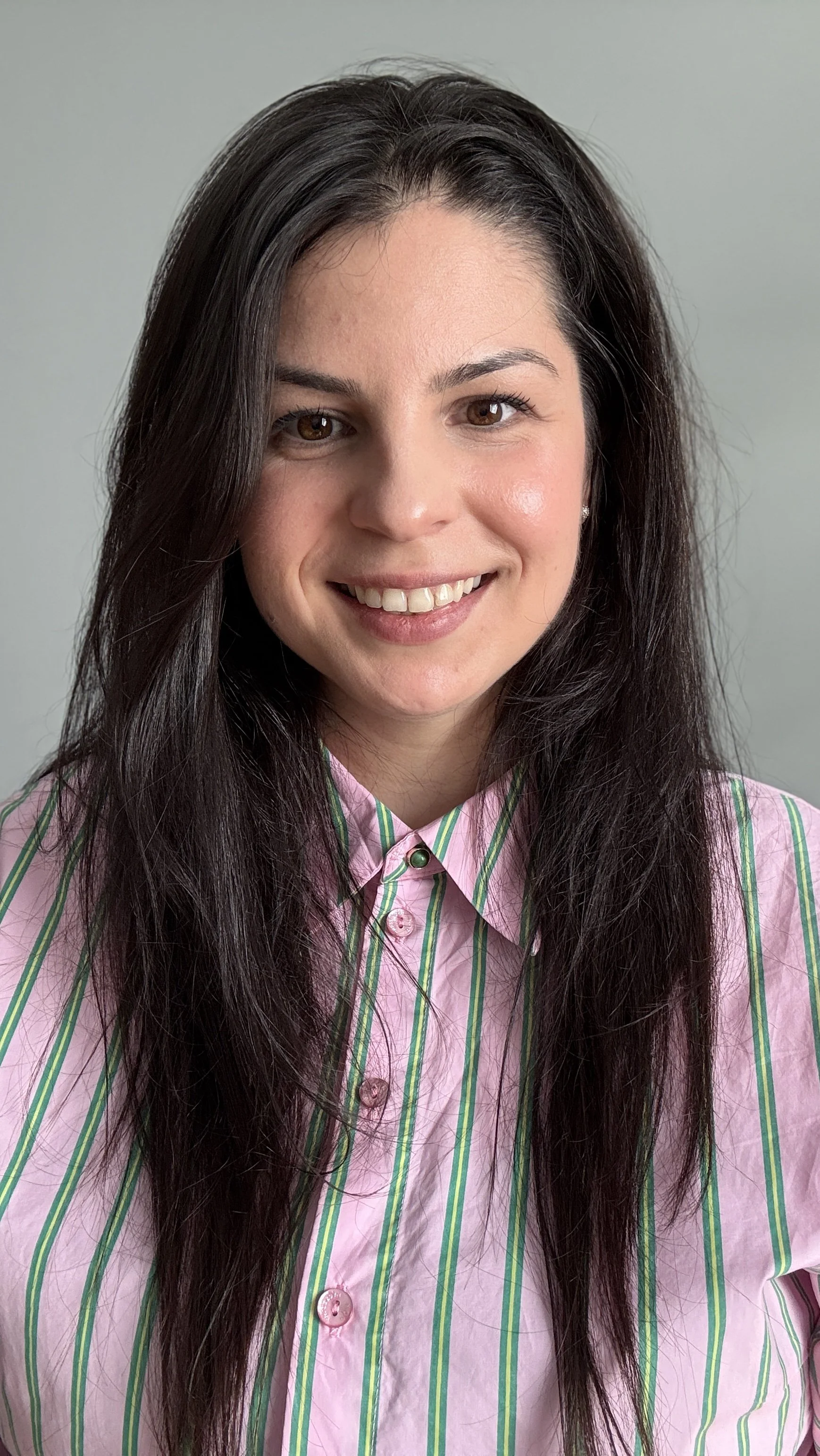 A woman with long dark hair, wearing a pink and green striped button-up shirt, smiling at the camera against a neutral background.