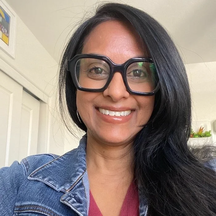 A woman with long black hair, wearing large glasses, a denim jacket, and a maroon shirt, smiling indoors.