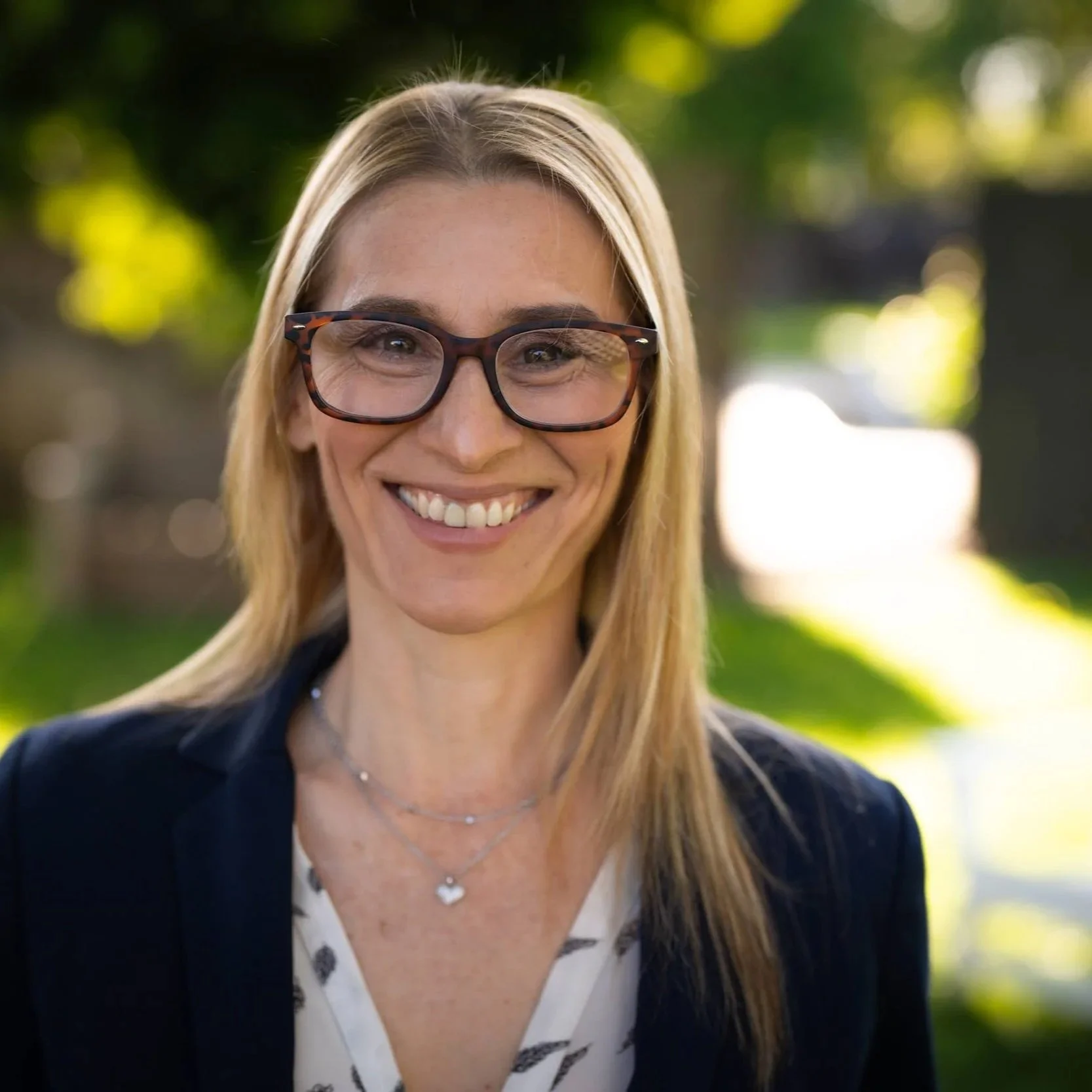 A woman with blonde hair, wearing glasses, smiling outdoors with a blurred green background.