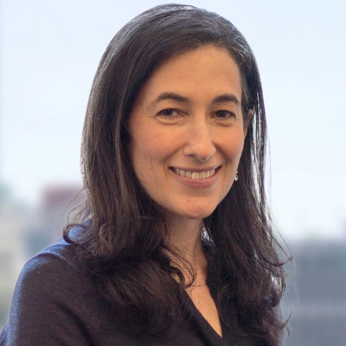 Portrait of a woman with long dark hair smiling, indoors with a blurred cityscape view in the background.
