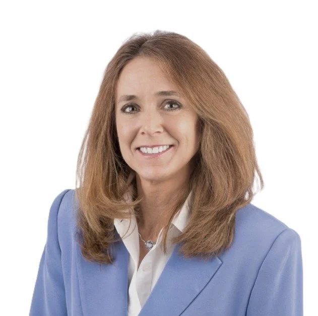 A woman with long, wavy, light brown hair wearing a blue blazer and white shirt, smiling against a plain white background.