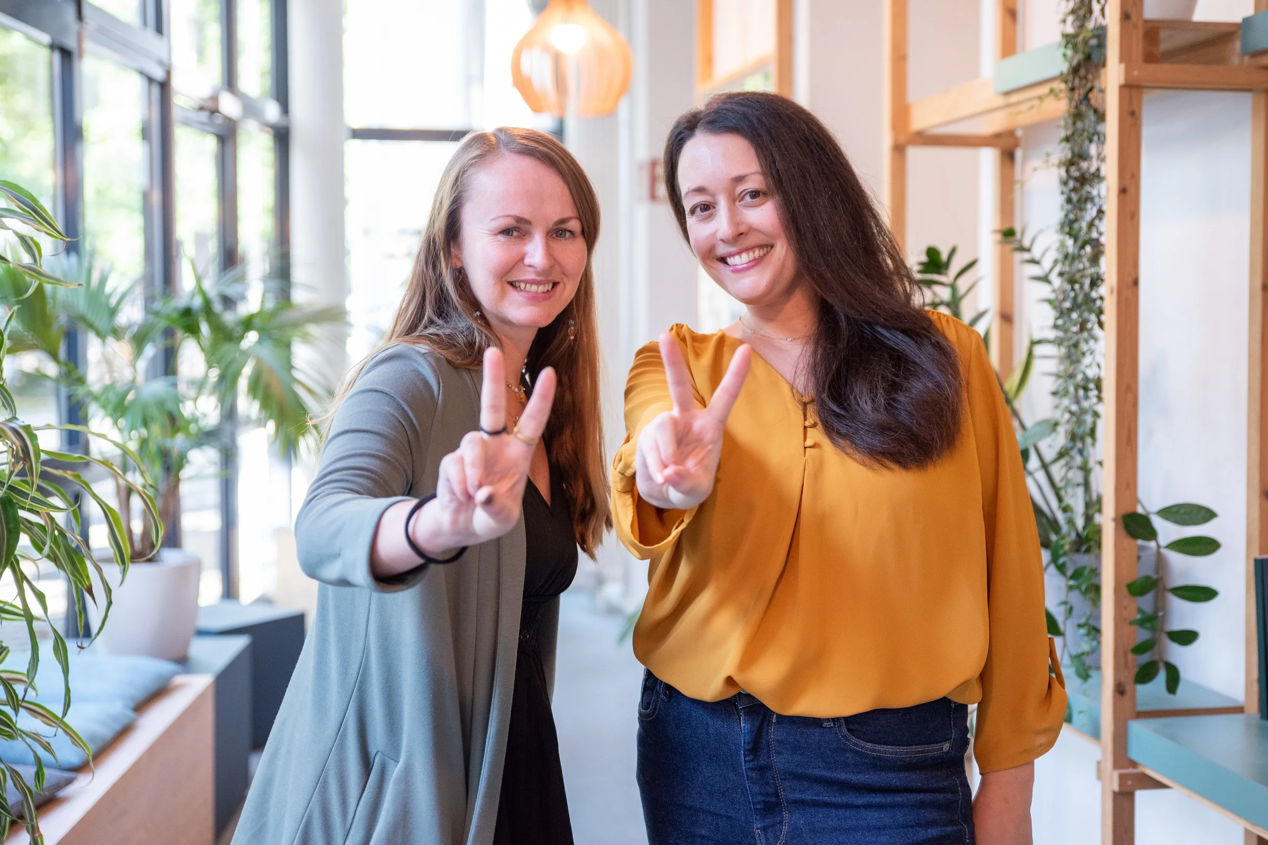 Two women smiling and making peace signs with their hands indoors with plants and wooden shelves in the background.