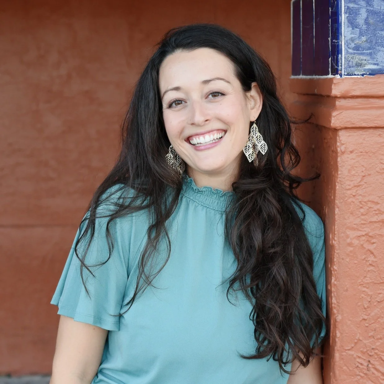 A woman with long dark hair smiling and wearing large leaf-shaped earrings and a teal blouse, standing next to an orange wall with blue decorative tile.