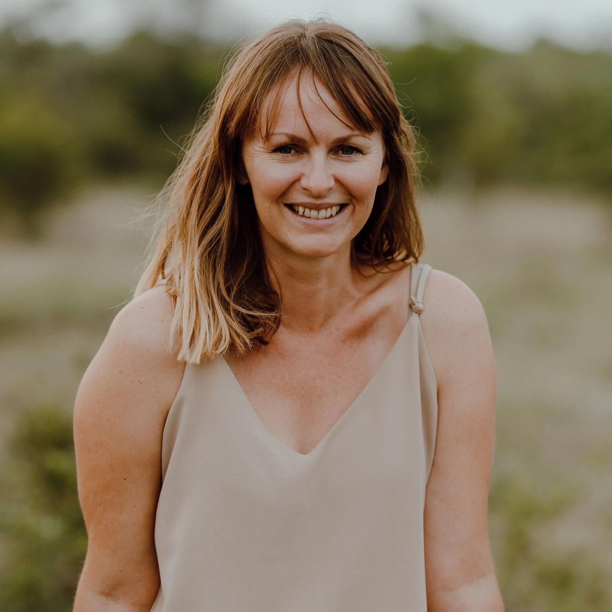 A smiling woman with shoulder-length light brown hair, wearing a beige tank top, stands outdoors with a blurred green landscape background.