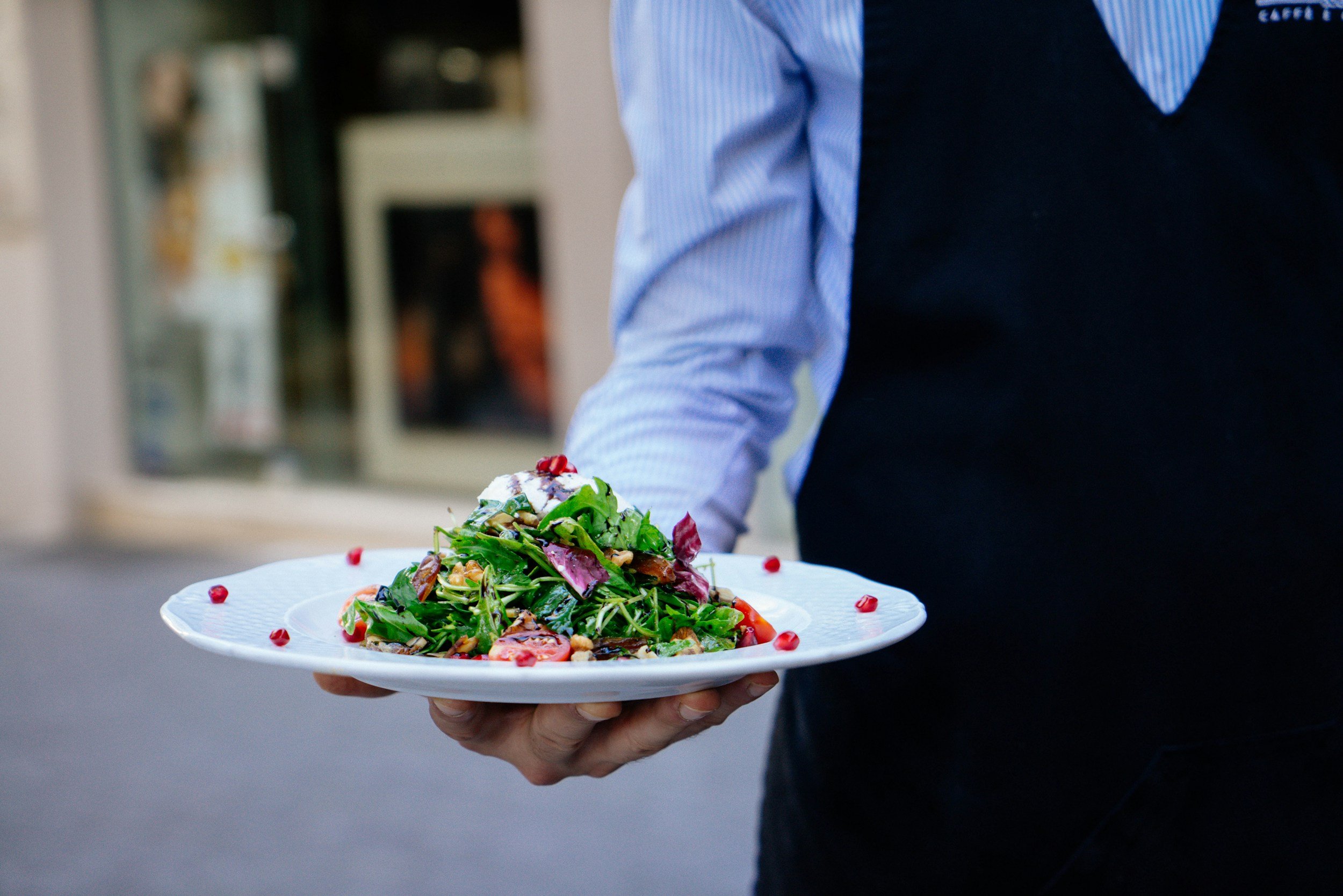 Person in a blue striped shirt and black vest holding a white plate of salad with greens, cherry tomatoes, and toppings outside a building.