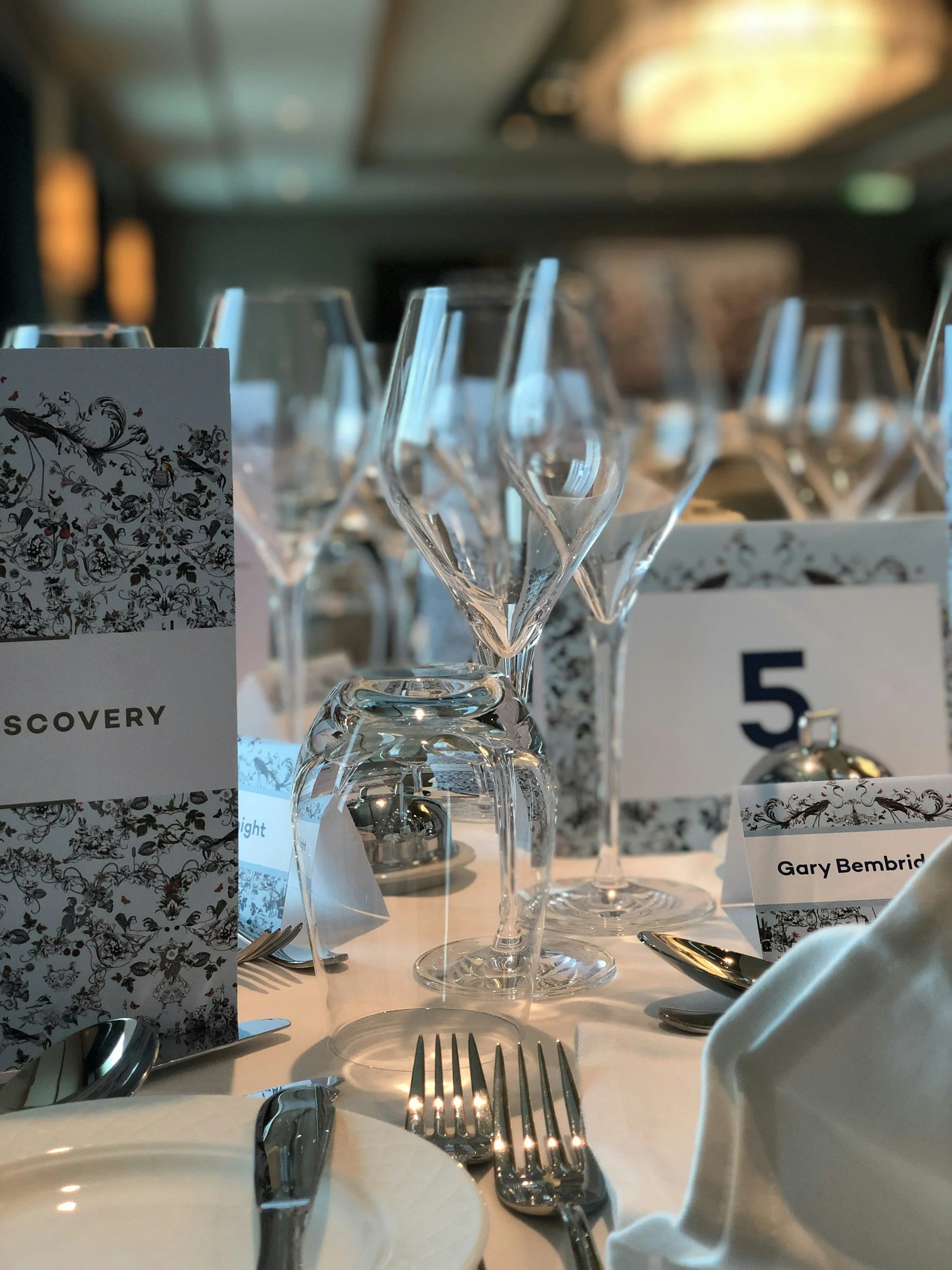 Table set for a formal event with wine glasses, silverware, numbered place cards, and decorative menus, in a dimly lit room.