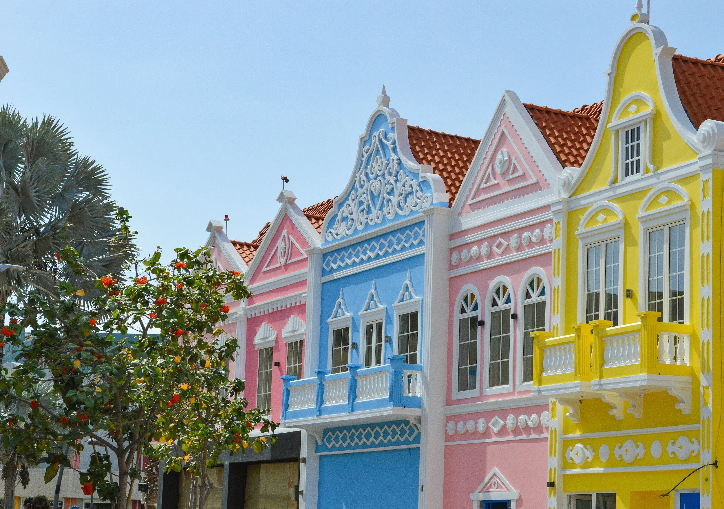 Colorful Victorian-style houses painted in pink, blue, and yellow with decorative details, next to trees and a clear blue sky.