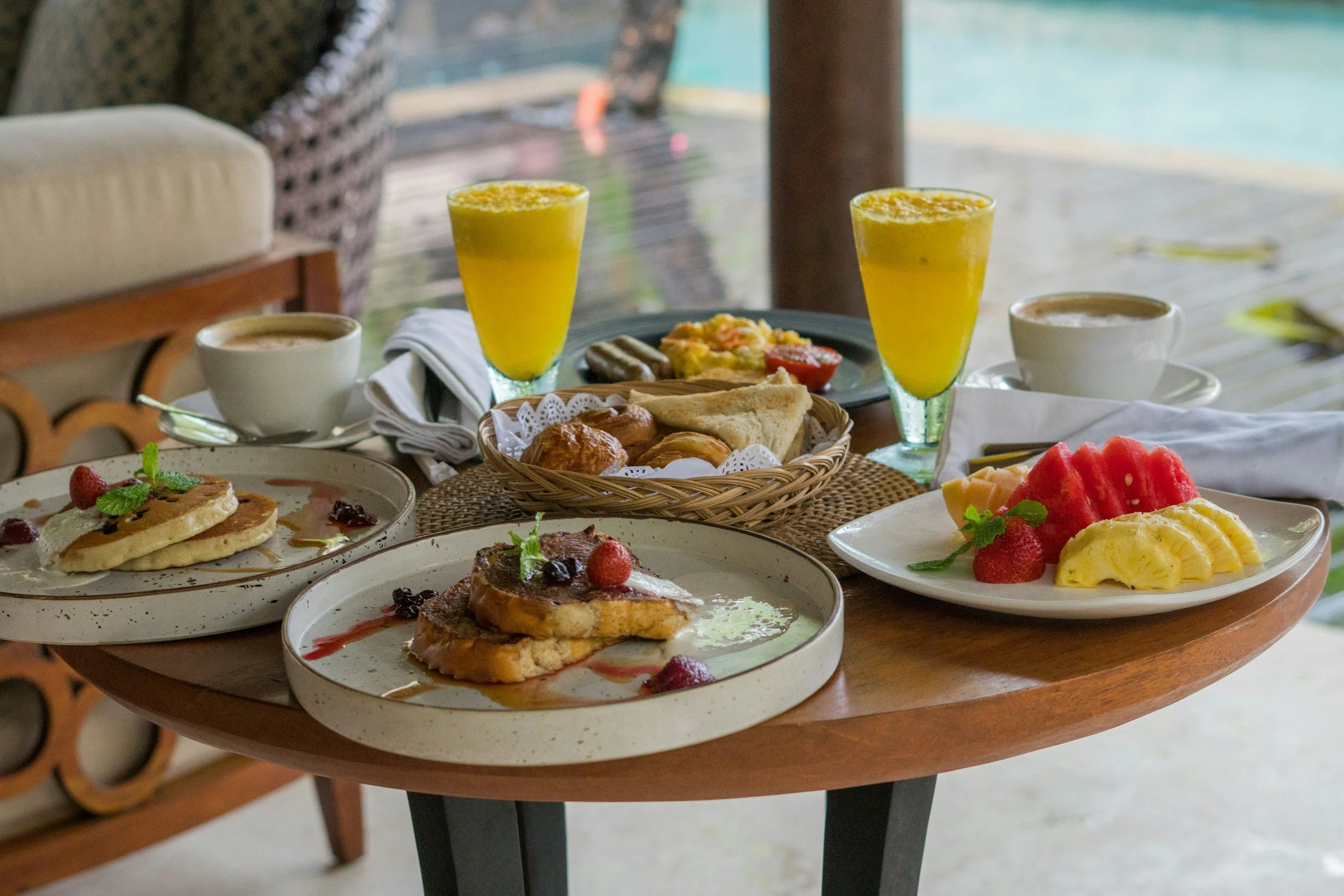 Breakfast spread featuring pancakes, French toast, fresh fruit, pastries, two glasses of orange juice, and cups of coffee on a wooden table near a body of water.