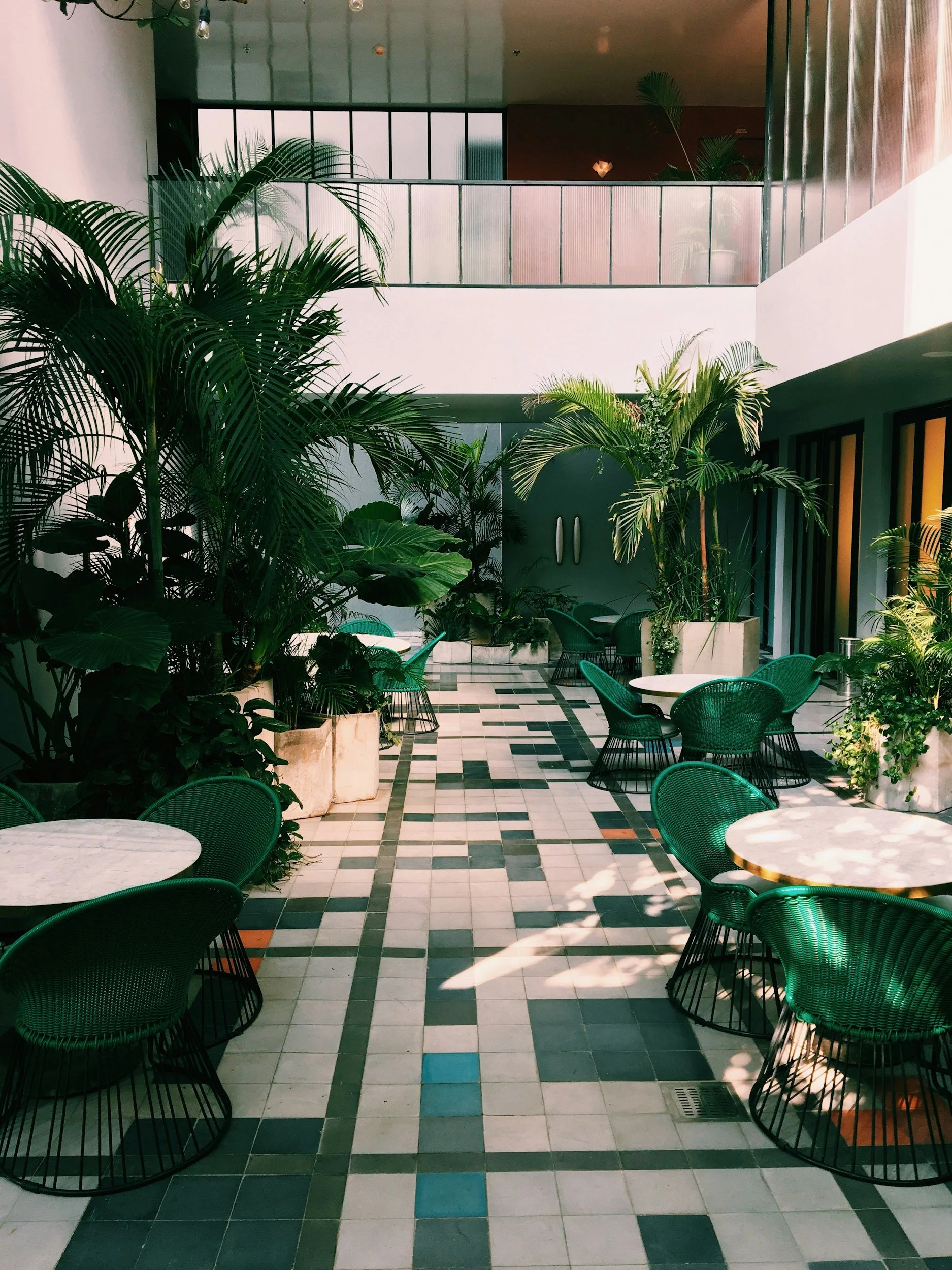 Indoor patio with green plants, round tables with white marble tops, and green wicker chairs. Sunlight filters through windows on the right, casting shadows on the tiled floor with a geometric pattern. Balcony railing visible on the upper level.