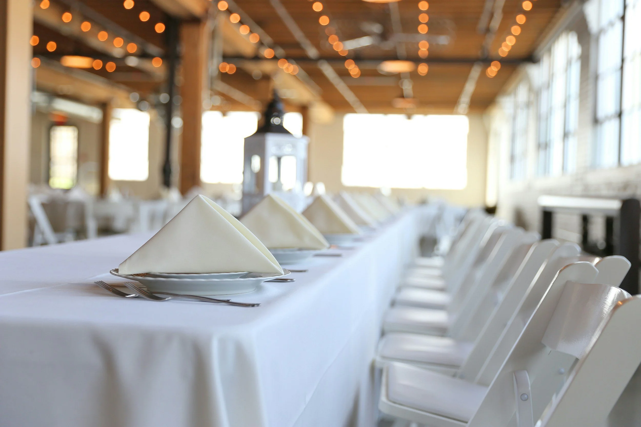 A long banquet table set for a formal event with white tablecloths, neatly folded napkins, and silverware in a spacious, rustic-style room with large windows and string lights.