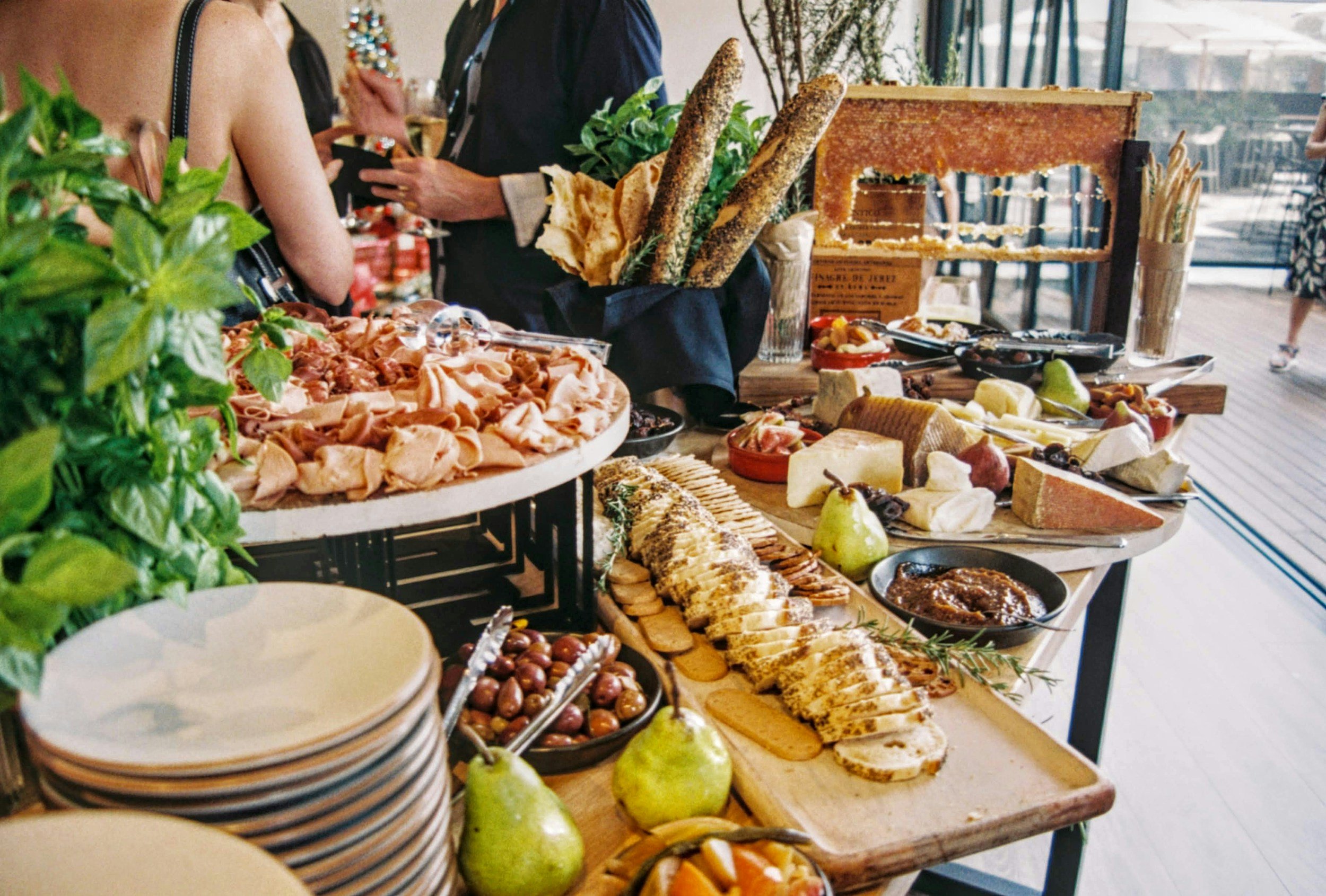 A buffet table filled with various cheeses, fruits, bread, and charcuterie, with people serving themselves in the background.