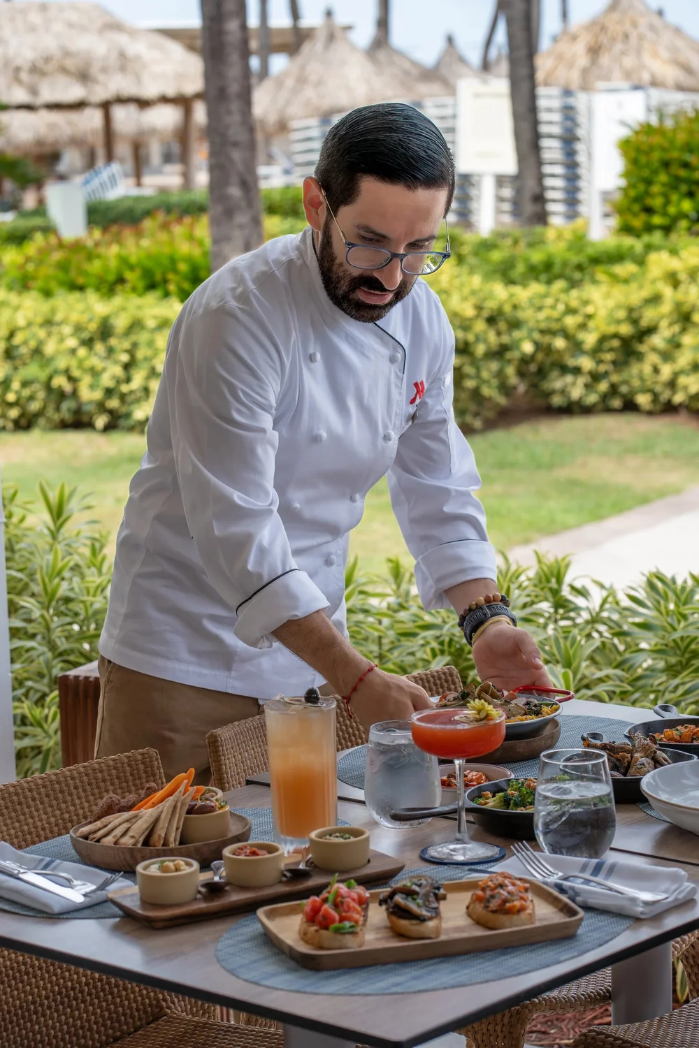 A chef in a white uniform preparing food at a table outdoors with tropical plants and thatched umbrellas in the background.
