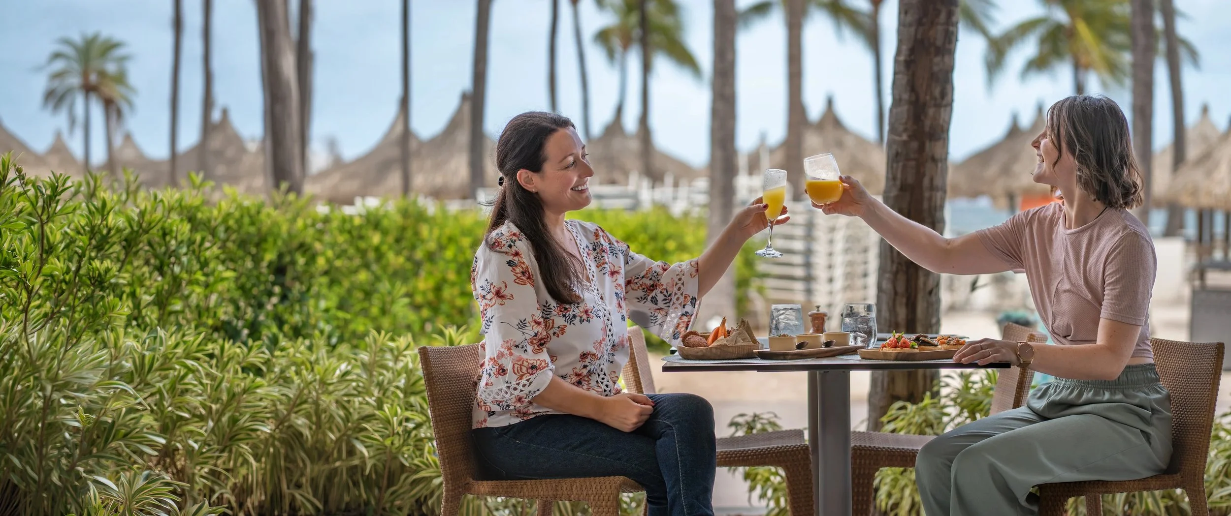 Two women enjoying drinks and food at an outdoor restaurant near palm trees and beach huts.