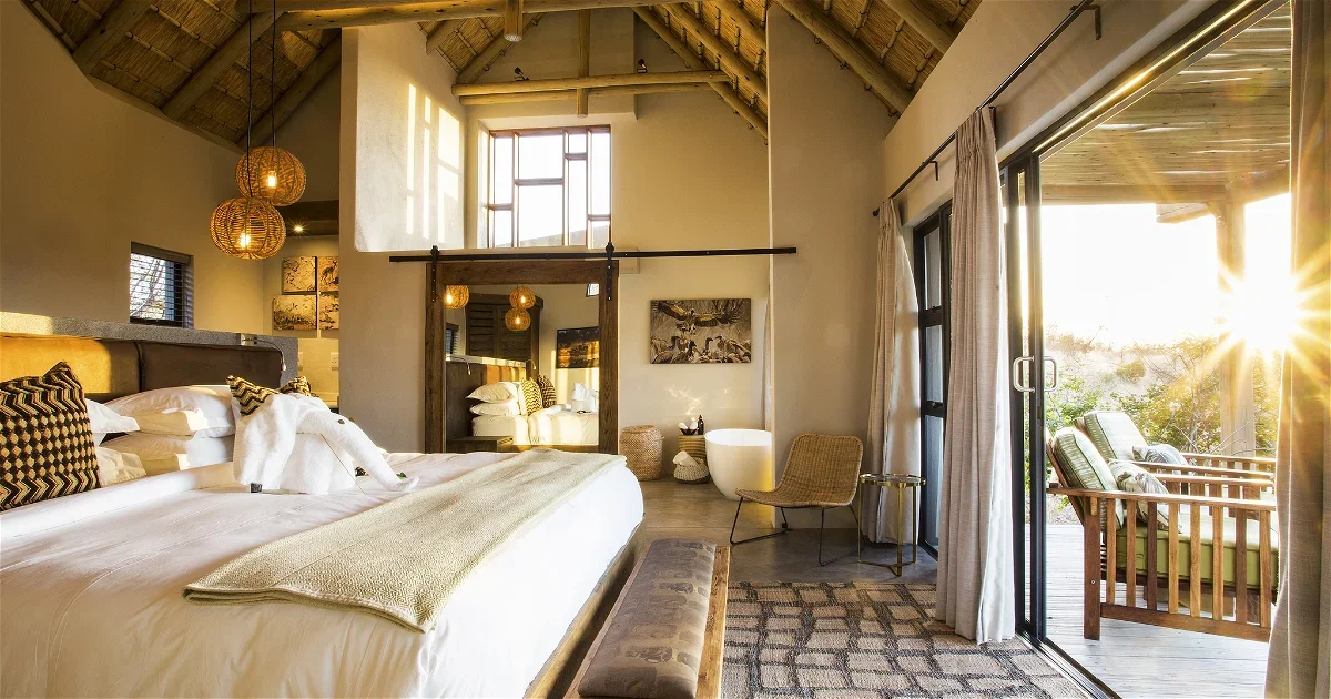 A bedroom with a large bed, decorative pillows, woven pendant lights, and a sitting area with a chair and sliding glass door leading to a balcony with outdoor seating, sunlight streaming in.