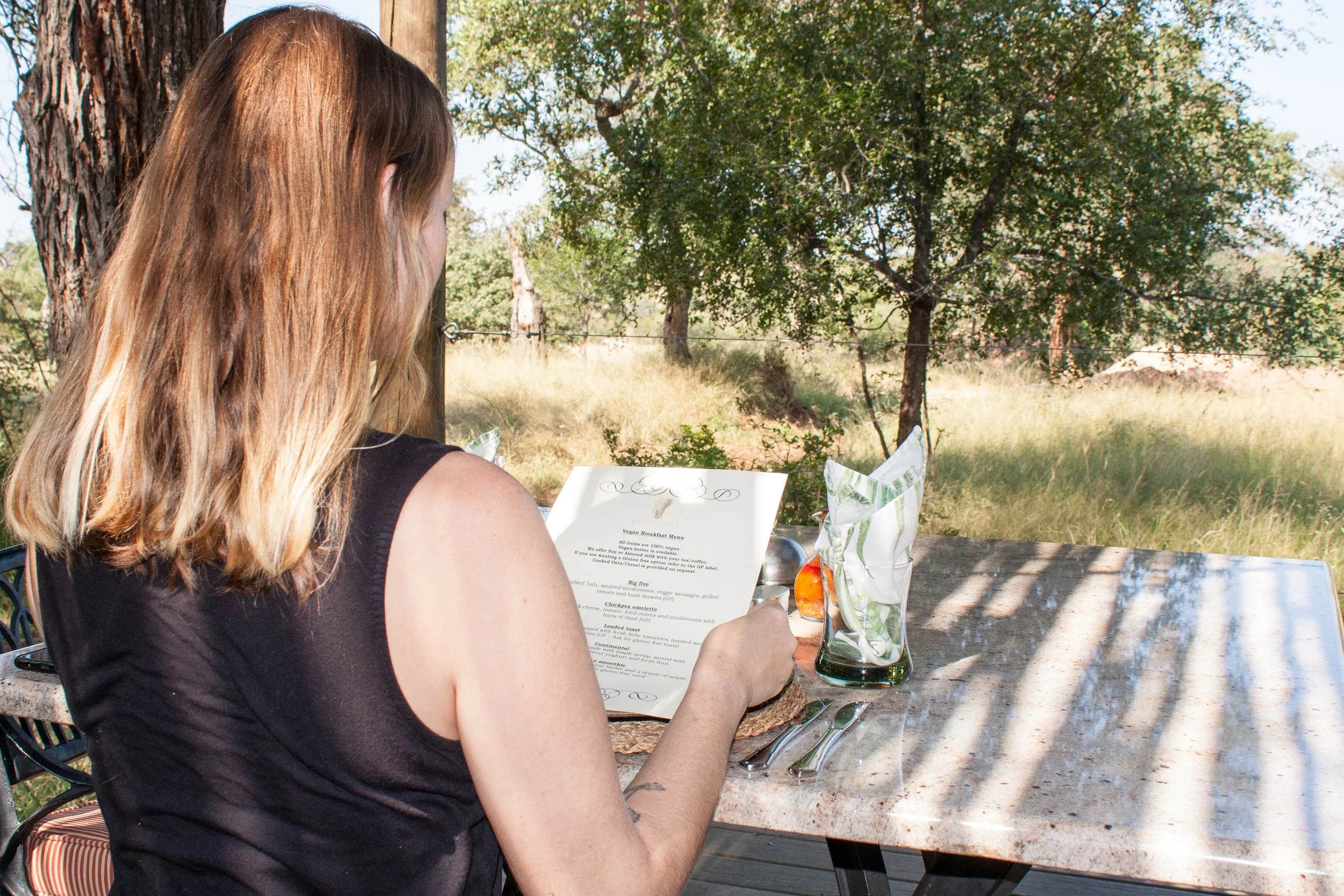 A woman sitting at an outdoor restaurant table, reading a menu with a scenic view of trees and grass in the background.