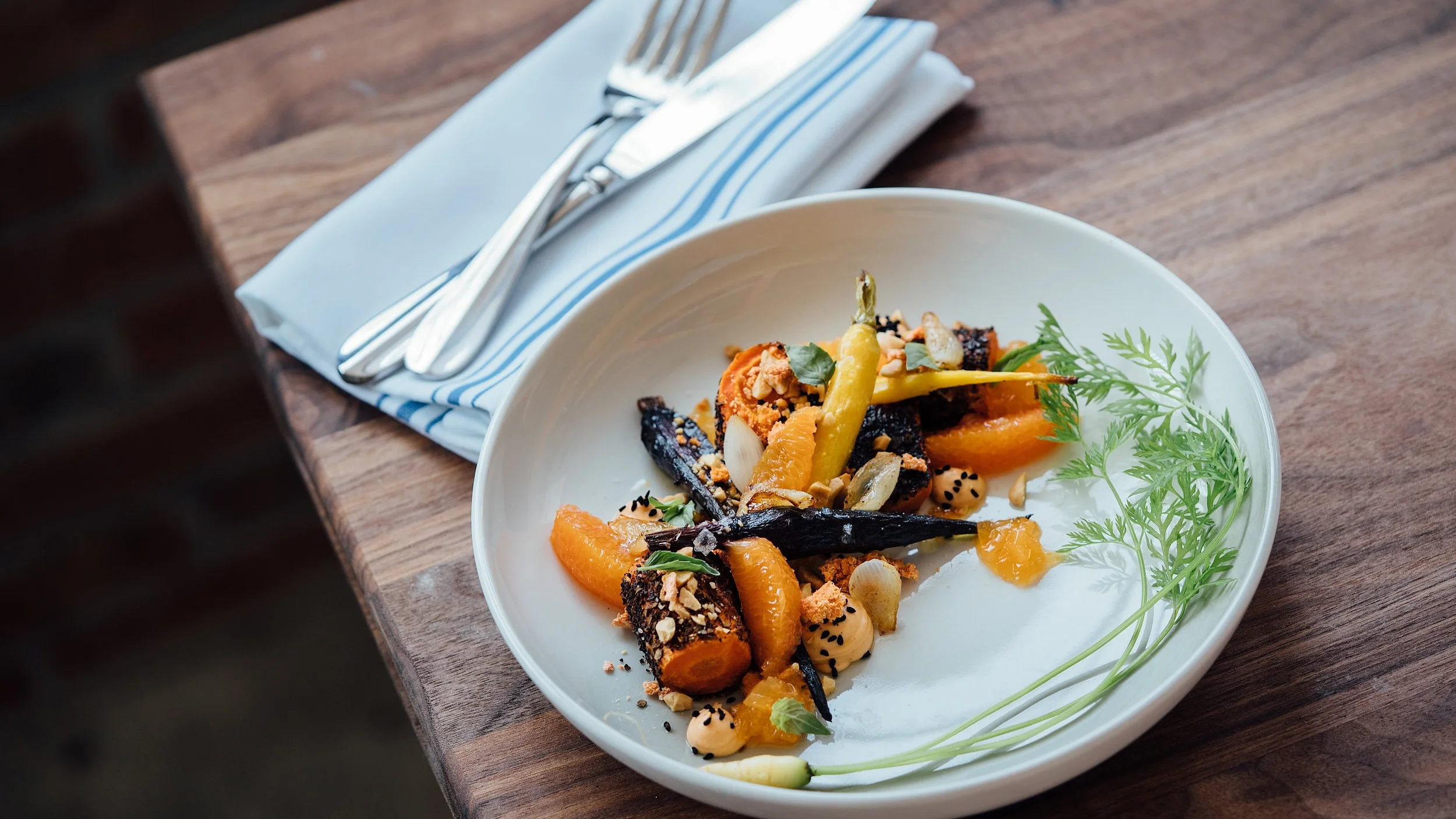 A white plate with colorful roasted vegetables and garnishes, placed on a wooden table beside rolled-up napkin with utensils.