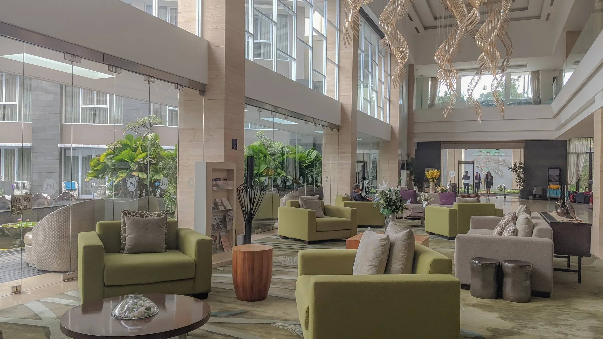 Modern hotel lobby with green and beige seating areas, large floral arrangements, and floor-to-ceiling windows showing outdoor greenery.