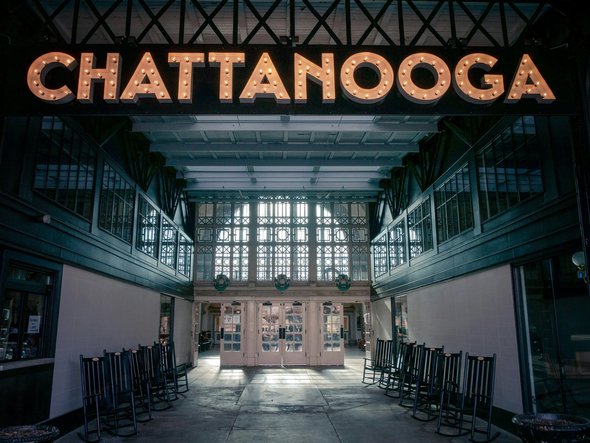 Interior view of Chattanooga train station with large illuminated sign reading 'Chattanooga' at the entrance, rows of empty chairs along the sides, and large windows letting in natural light.