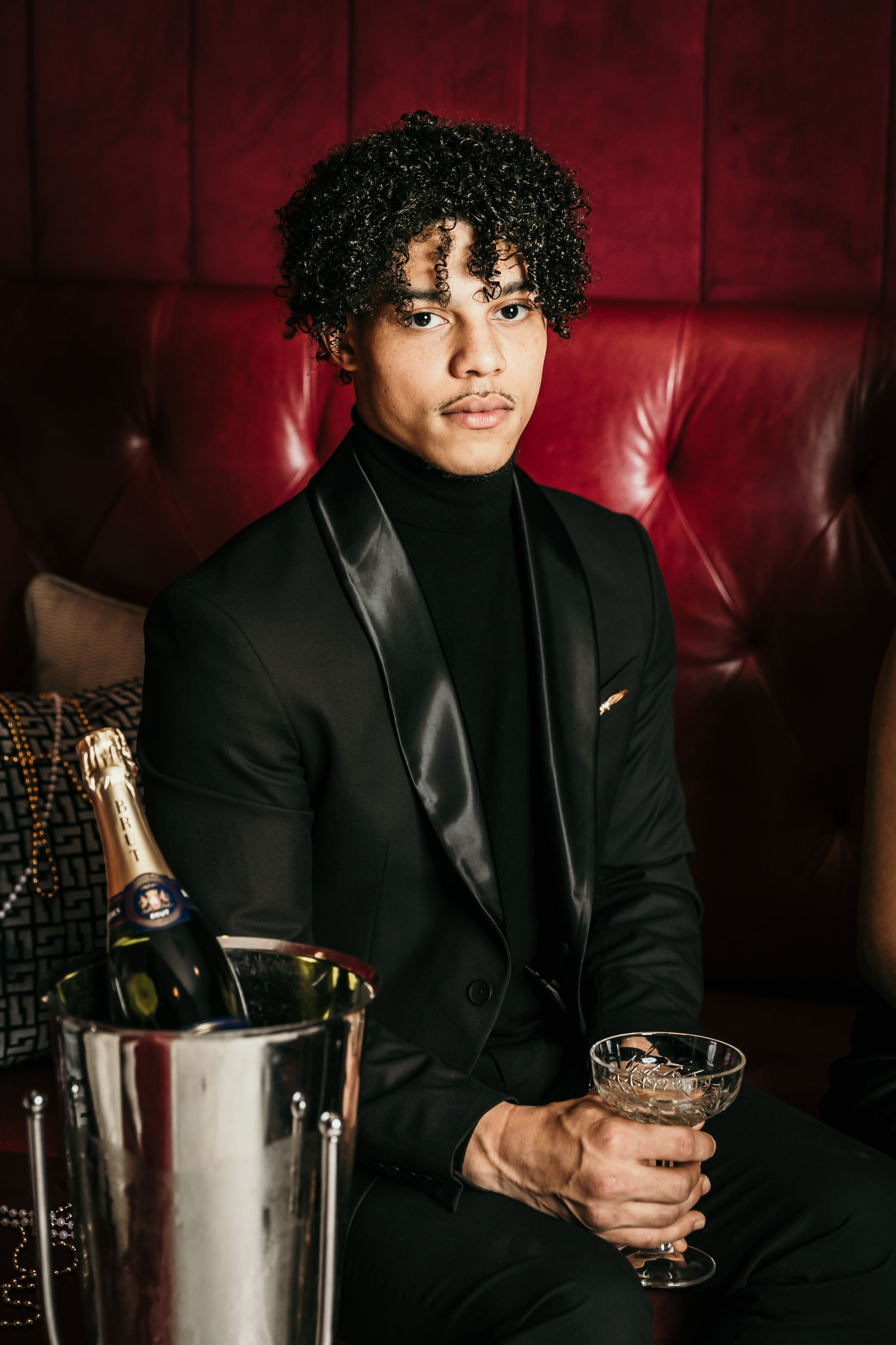 A young man with curly hair in a black suit and turtleneck, holding a glass of drink, sitting on a red leather couch at a celebration, with a bottle of champagne in an ice bucket nearby.