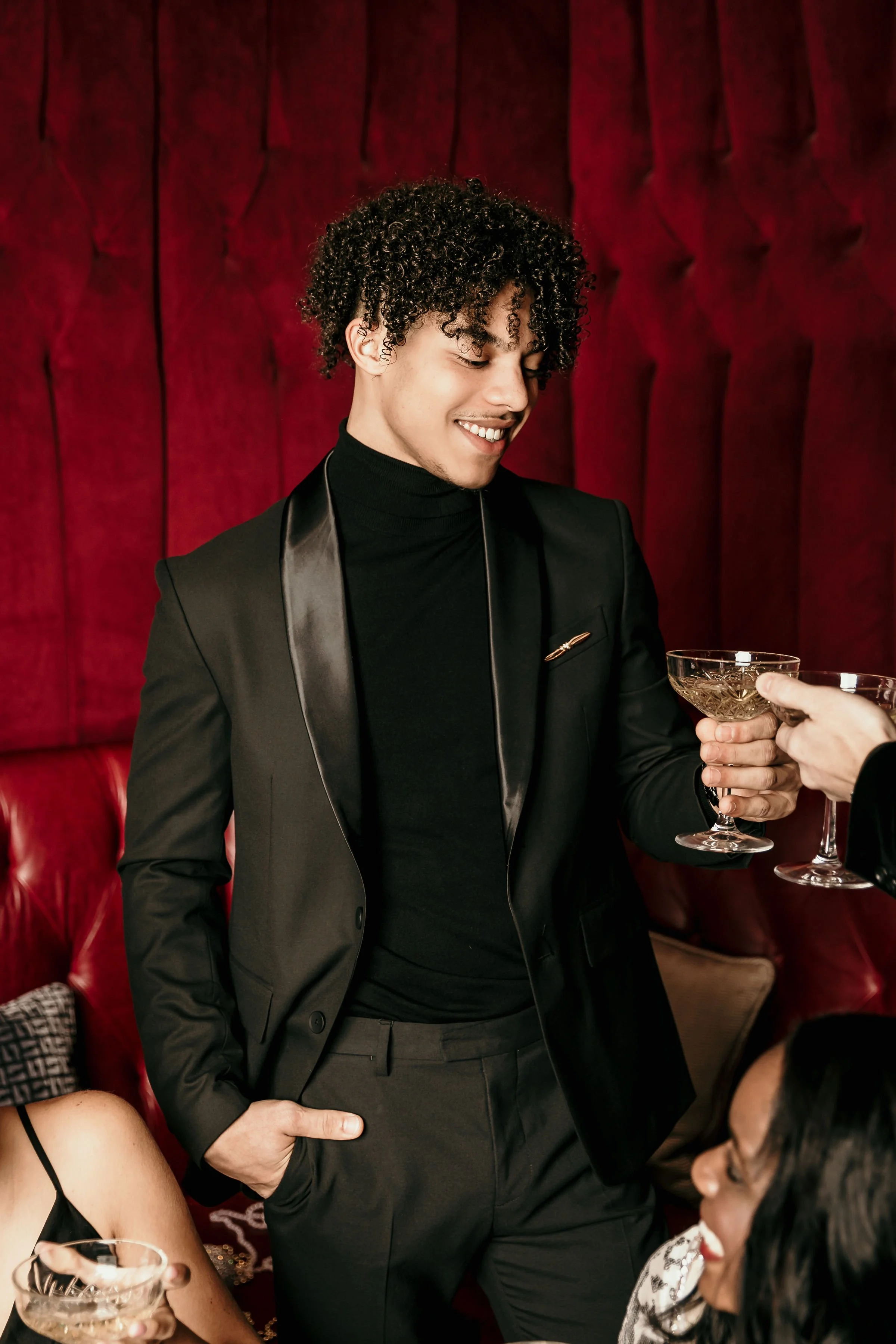 A man dressed in a black tuxedo with satin lapels, black turtleneck, is smiling and holding a champagne coupe glass. He is at a celebration or party, with a woman sitting near him, also holding a glass, in a setting with red velvet walls.