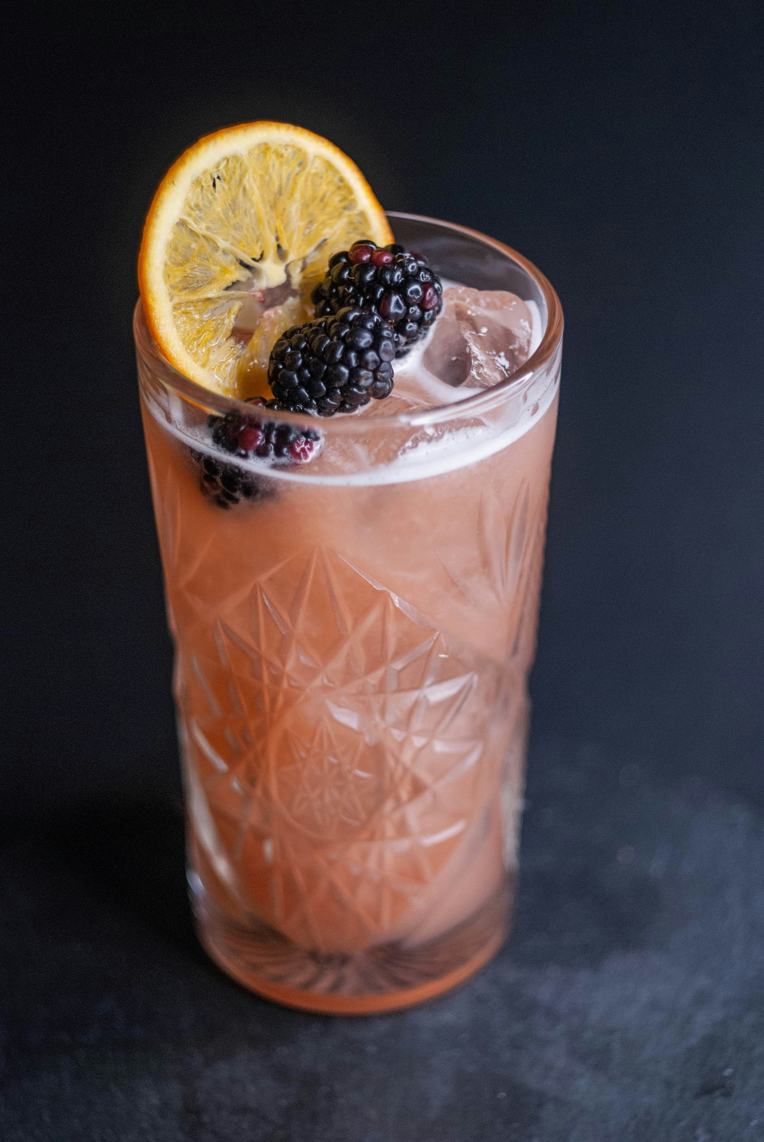 A tall glass of pink beverage with ice, garnished with a slice of orange and blackberries, on a dark surface.
