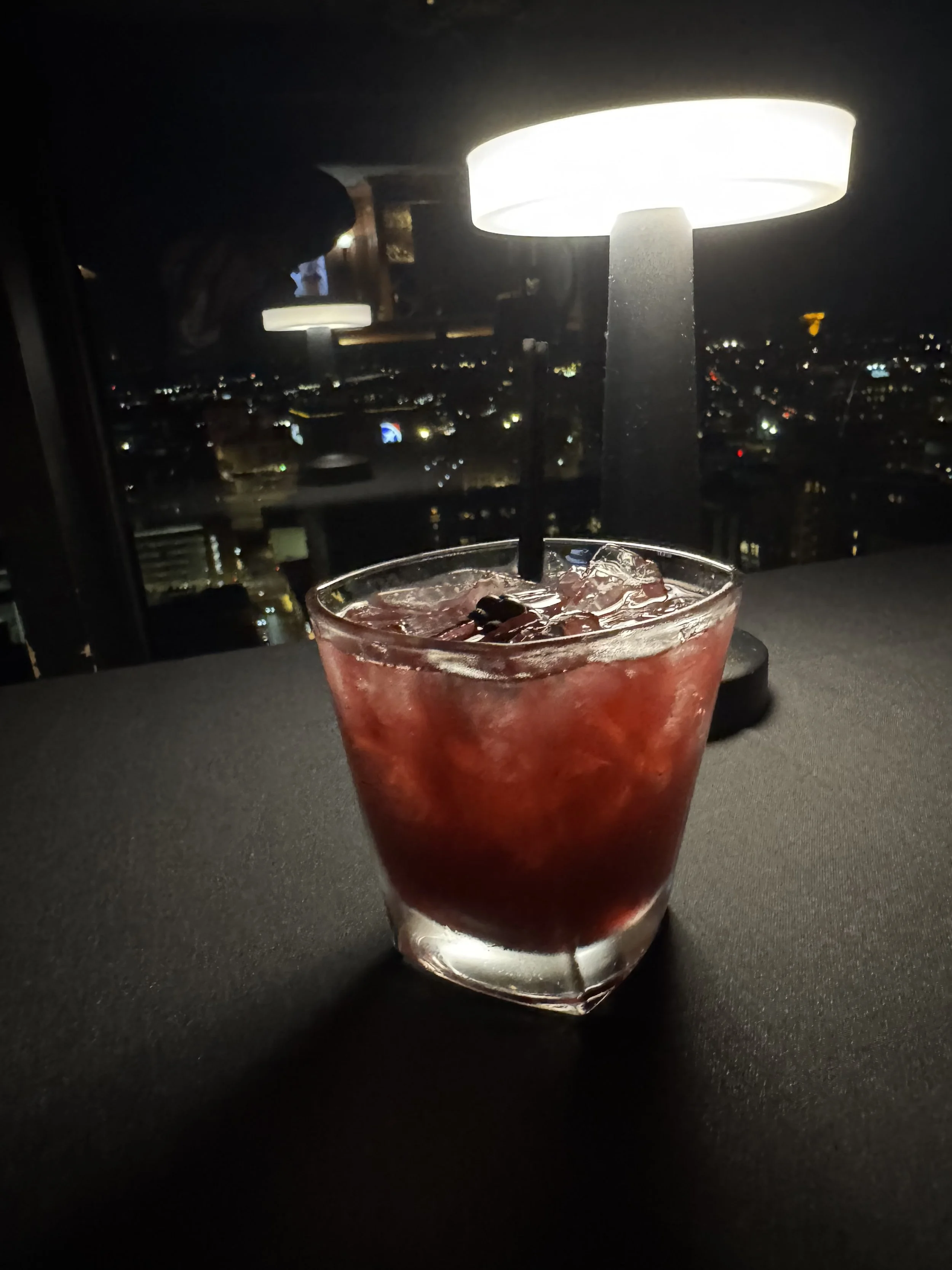 A glass of red cocktail with ice cubes and a straw on a dark table, with a city skyline and night lights in the background.