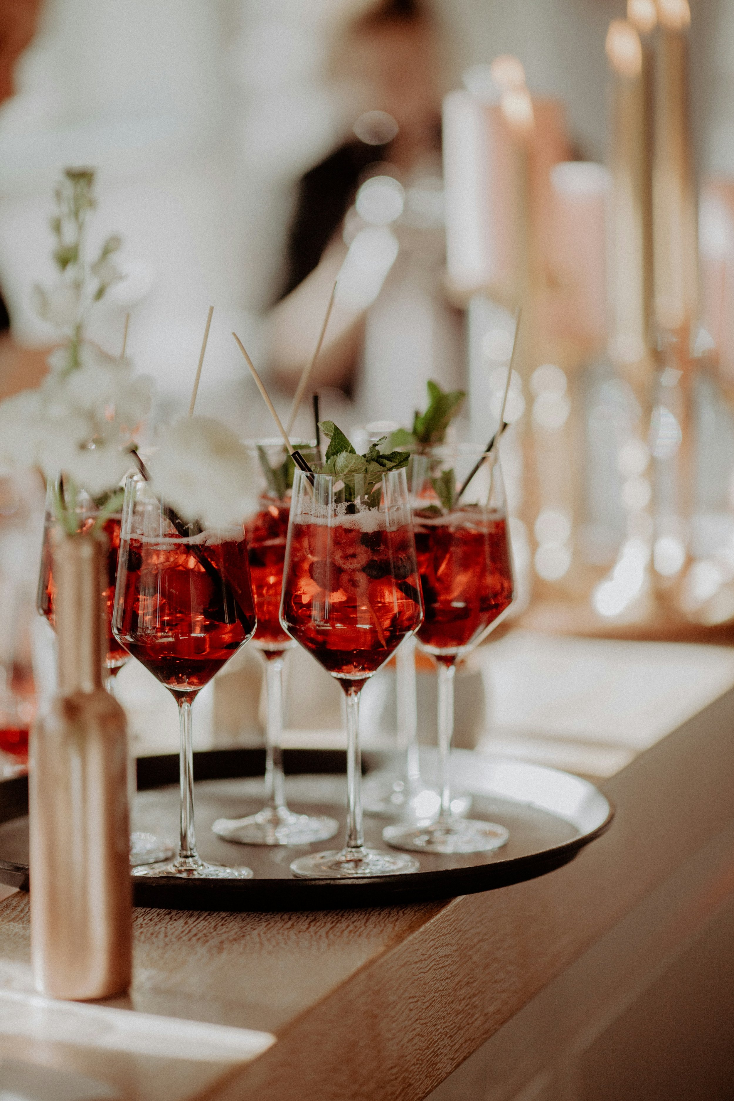 Several glasses of red cocktail drink garnished with mint leaves on a black tray, placed on a wooden surface, with blurred background of a person pouring drinks.