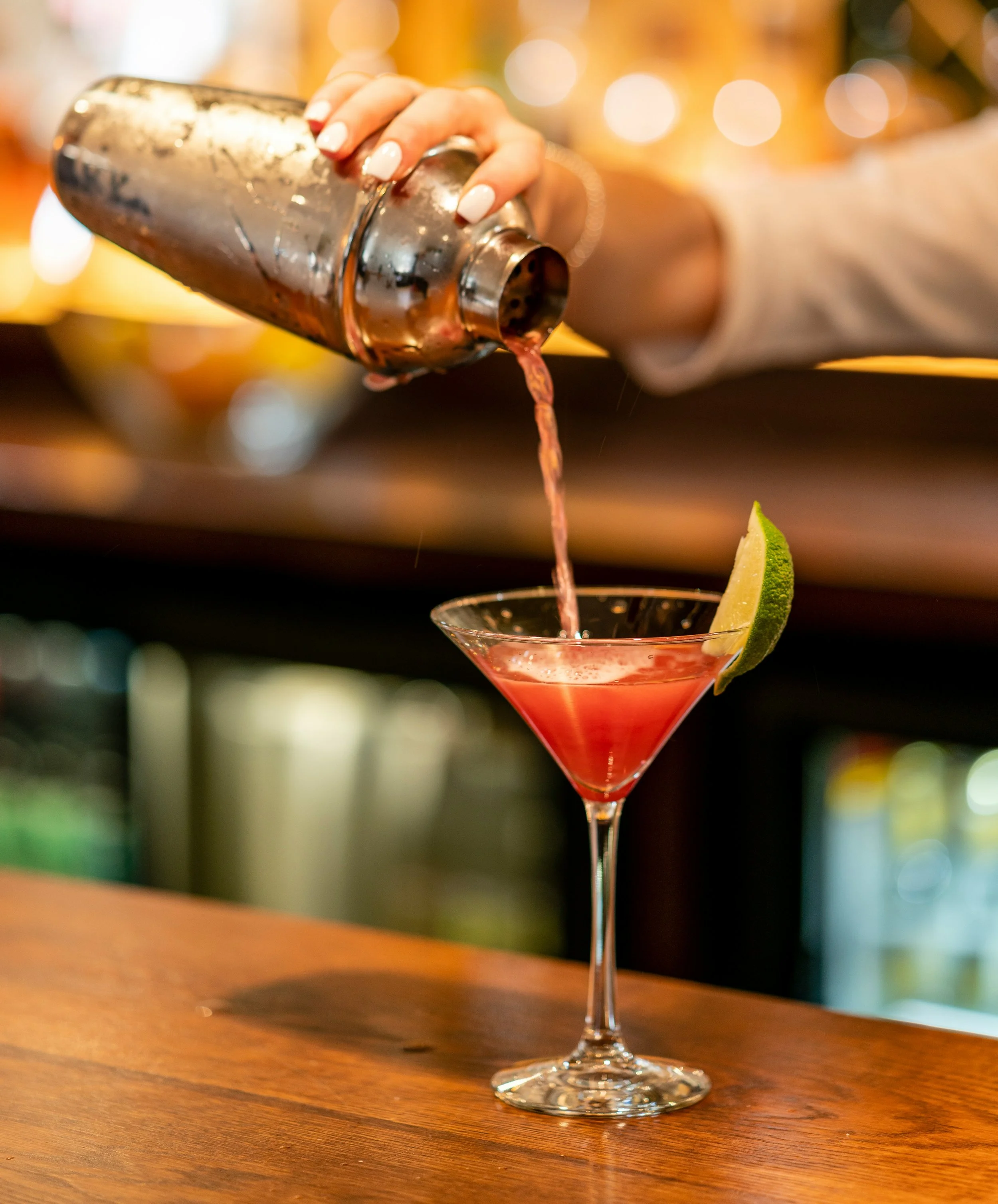 A bartender pouring a pink cocktail from a shaker into a martini glass garnished with a lime wedge on a wooden bar counter.