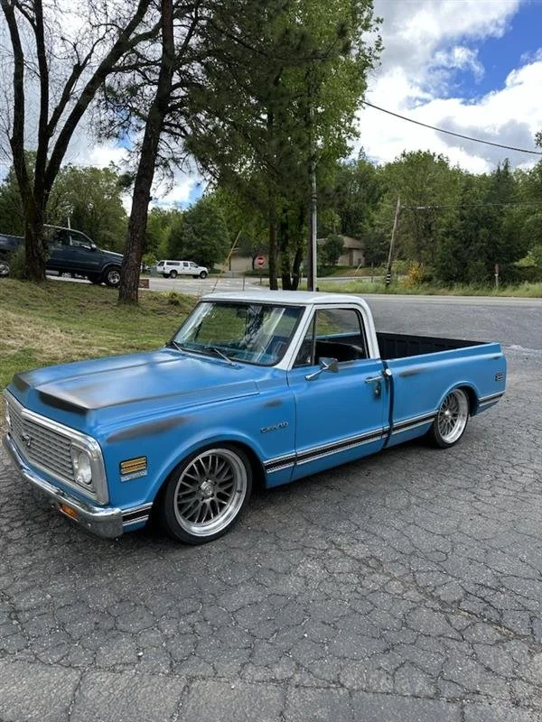 A vintage blue and white pickup truck parked on a cracked asphalt surface during the daytime with trees and a few cars in the background.