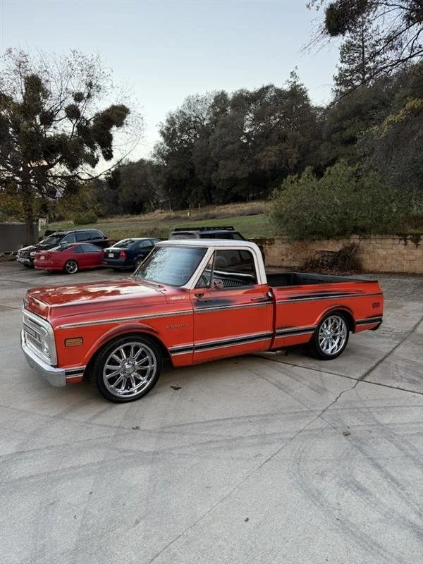 A vintage red pickup truck with chrome rims parked on a concrete surface, with other parked cars and trees in the background.